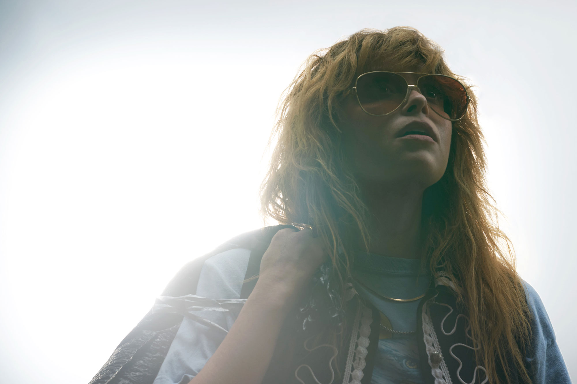 Image of Natasha Lyonne looking up and to the right, off camera, in an overexposed image so bright that she's the only thing visible. She is wearing aviator sunglasses and is holding her jacket over her right shoulder. 