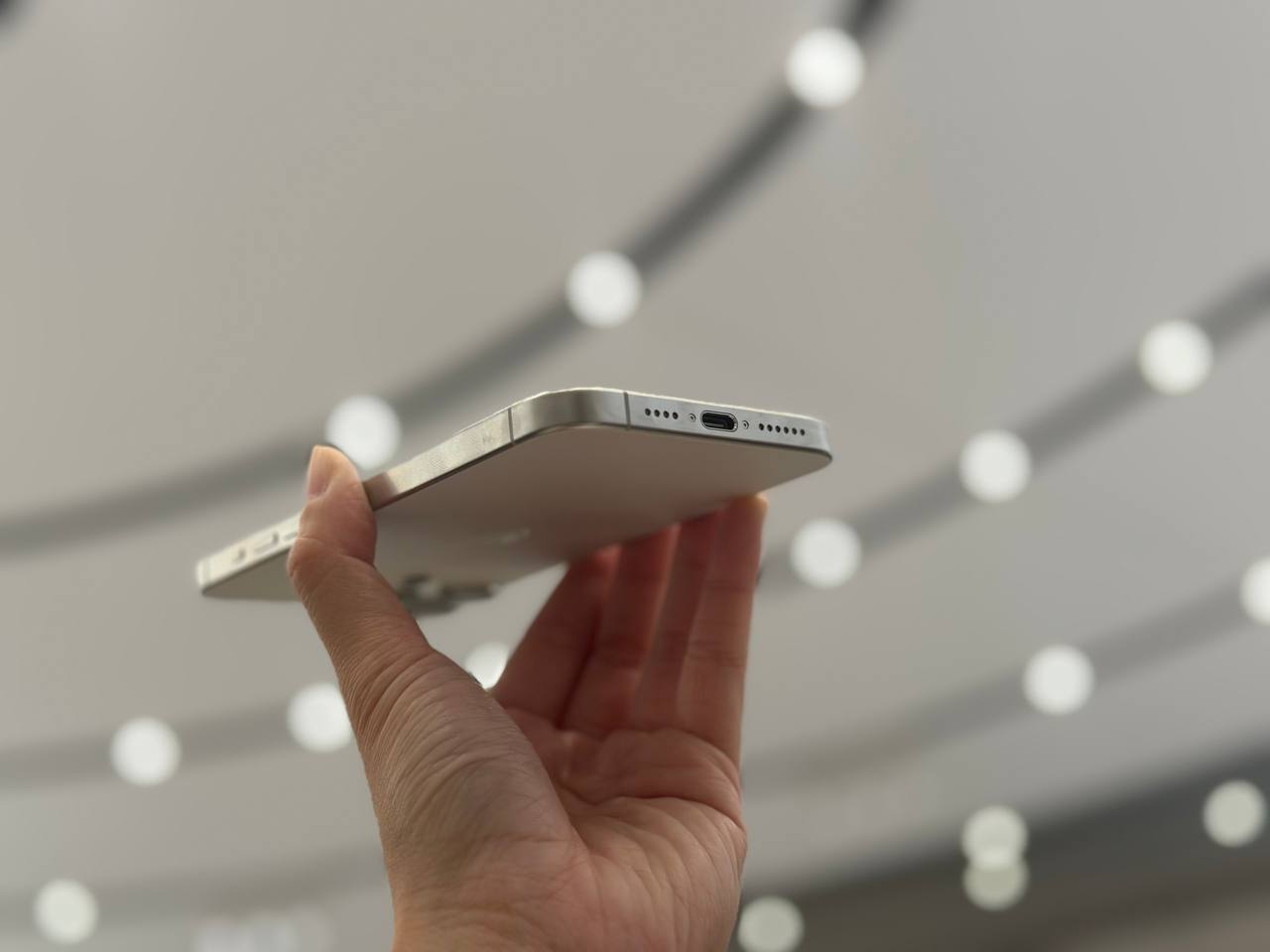 The iPhone 15 Pro Max held up against a ceiling of lights, showing its USB-C port.