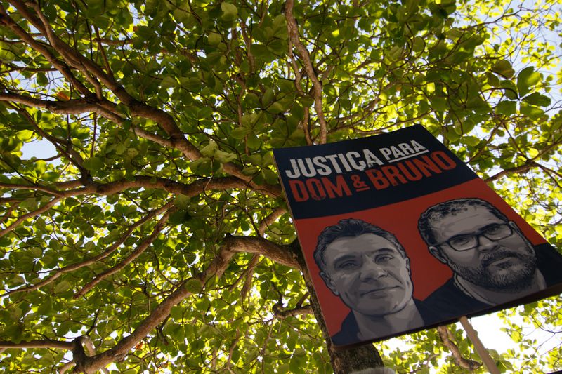 A sign saying “Justica Para Dom &amp; Bruno” is held in front of a tree.