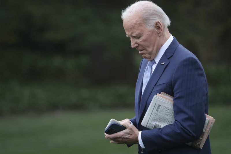 Biden looks at his cellphone while holing a newspaper under one arm.
