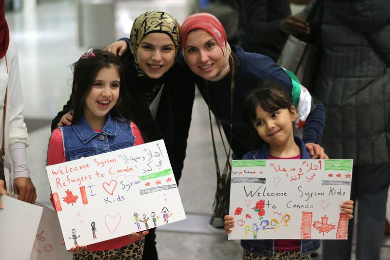 Two children and two women hold child-made illustrated signs welcoming Syrian refuges. One sign reads “Welcome Syrian refugees to Canada, we (heart) Syrian kids,” the other reads “Welcome Syrian kids to Canada.” Both signs are also written in Arabic.