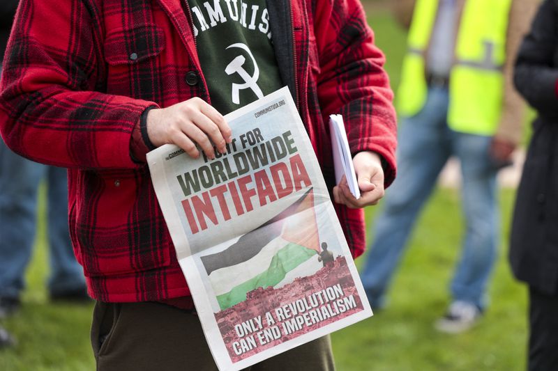A young man in a red plaid jacket and a T-shirt with a communist symbol holds a colorful printed newsletter reading “Worldwide Intifada,” among other text and images.