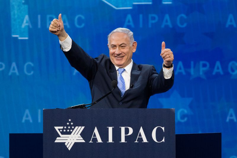 Benjamin Netanyahu, in a black suit, smiles and gives two thumbs up behind a podium labeled “AIPAC” on a stage. 