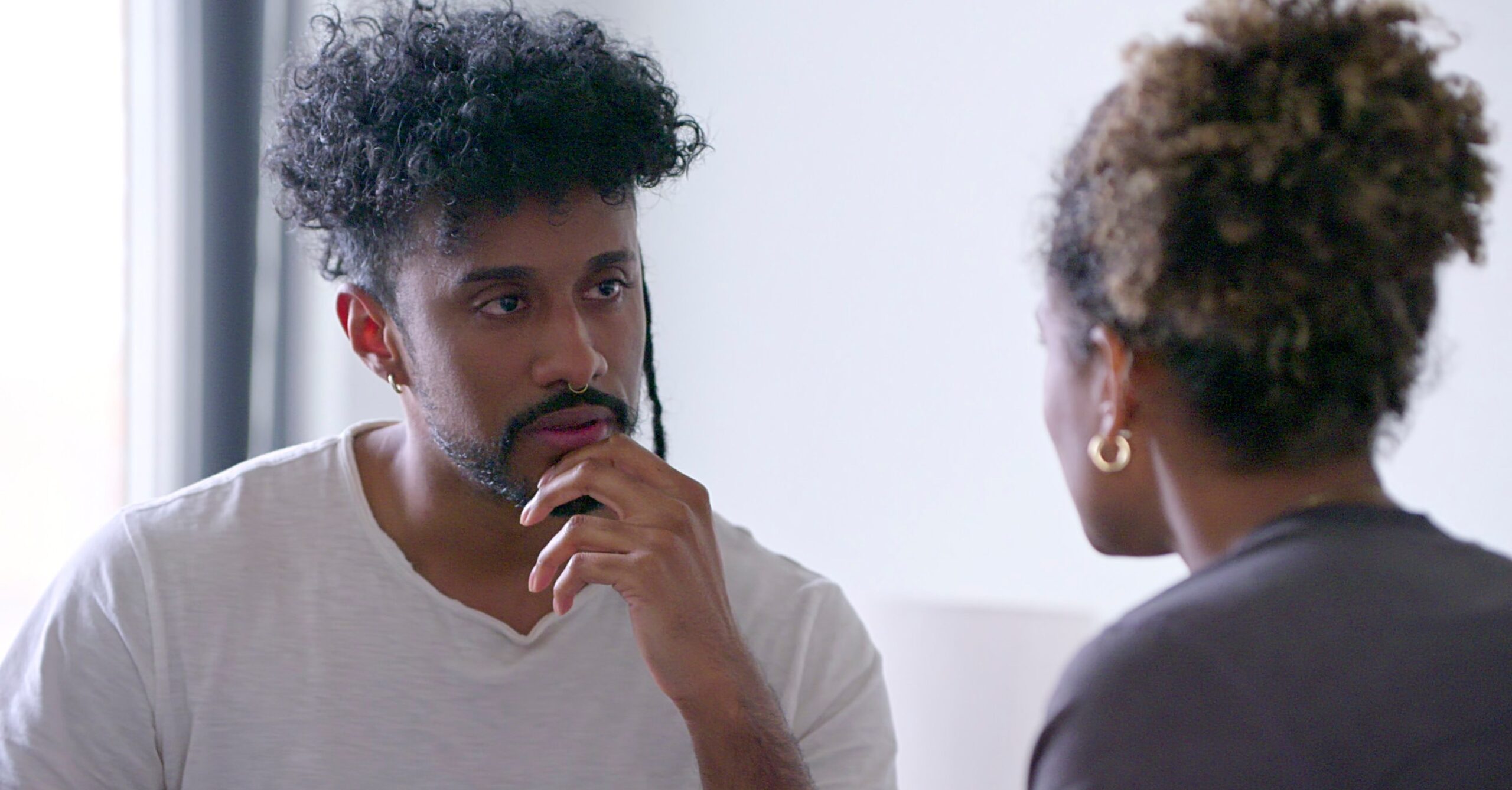 A man sits with his hand to his chin, speaking to a woman we see in profile.