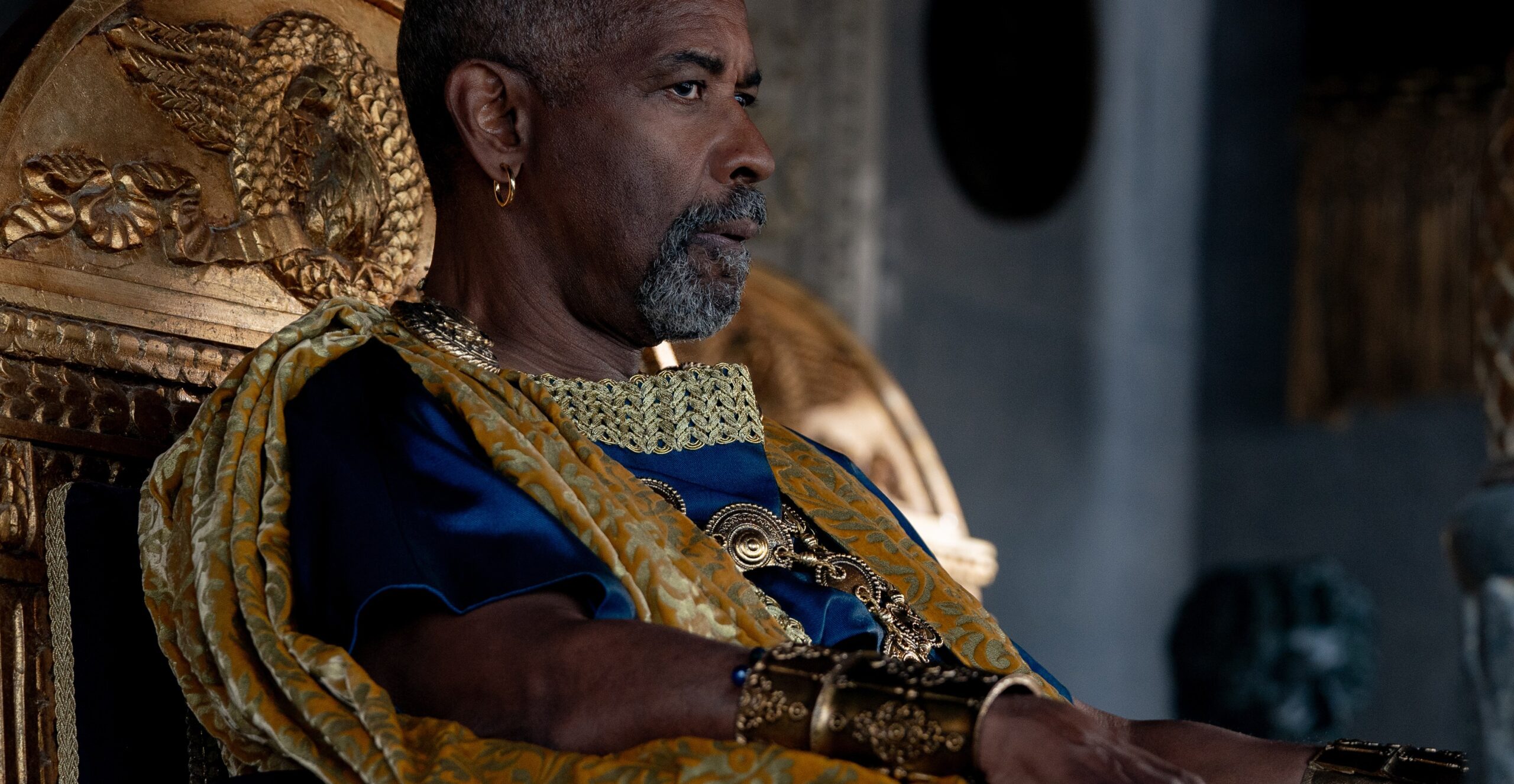 Denzel Washington in blue and gold Roman dress sitting on a golden throne-like chair.