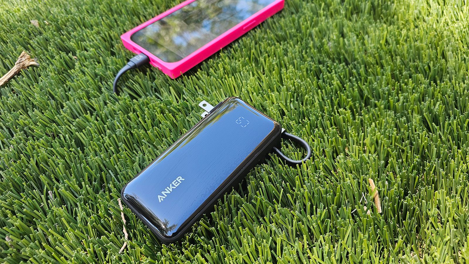 The Anker 10K Fusion power bank charges an iphone with its built in cable as both devices sit on some fake grass on a hot day. 