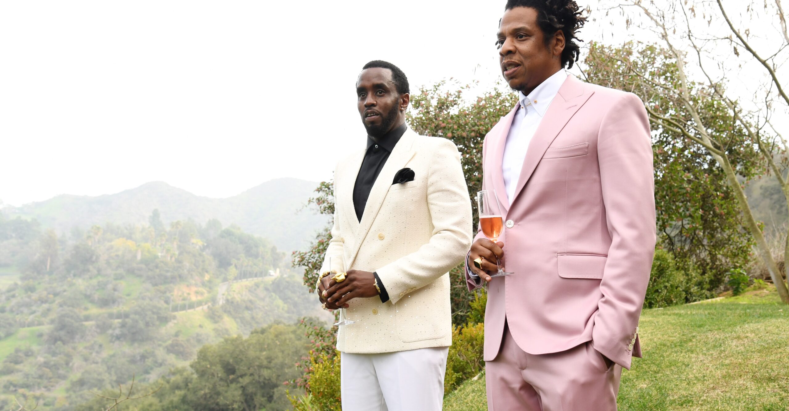 Diddy in a white suit and black shirt stands next to Jay-Z in a pink suit and white shirt. Both men are holding wine glasses and standing outdoors at the edge of a manicured lawn with a wild hillside behind them.