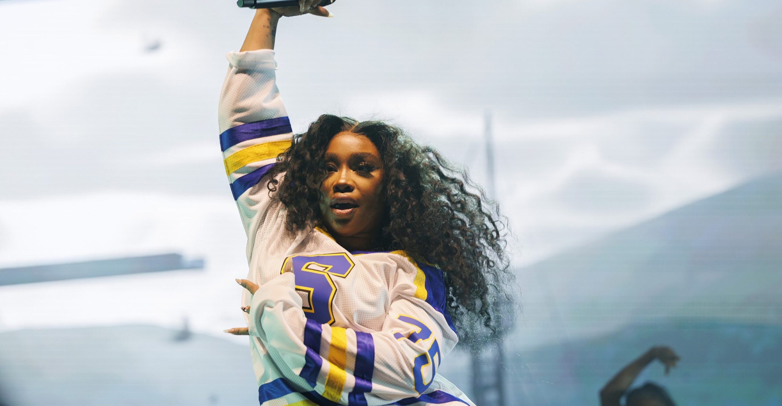 A young Black woman with long hair and wearing a white, blue, and yellow football jersey dances onstage with a microphone held over her head.