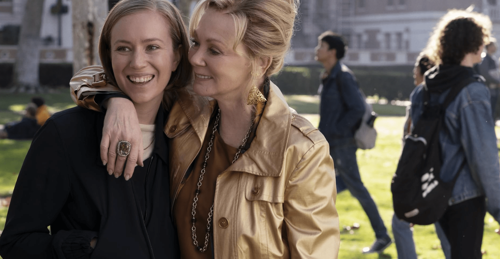 Two blond women in a park stand close and smile.