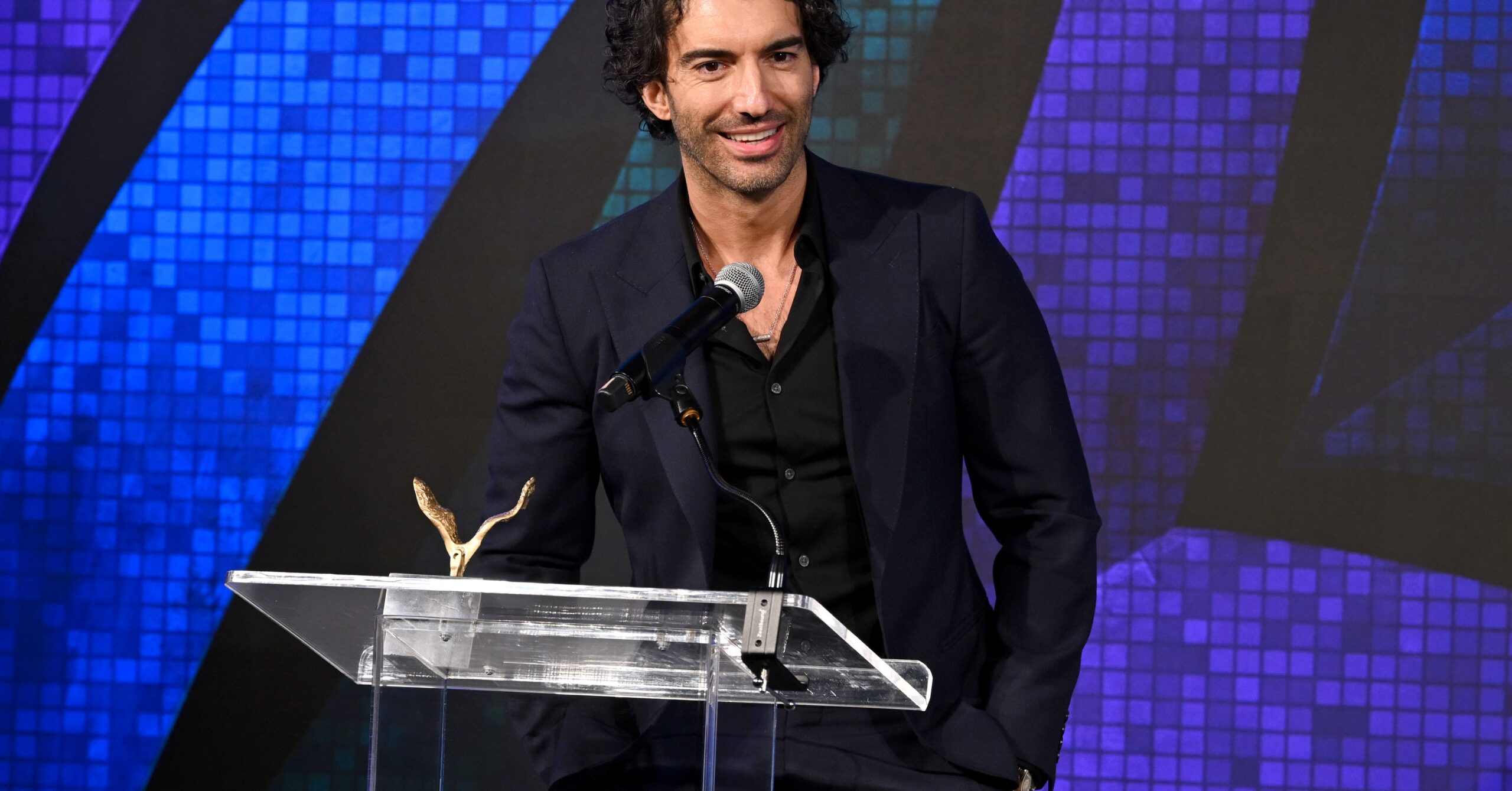 A man in a black suit standing onstage at a plexiglass lectern.