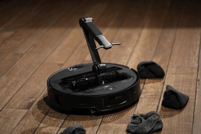 A black robot vacuum on a wooden floor surrounded by socks. A robotic arm is coming out of the center of the robot.