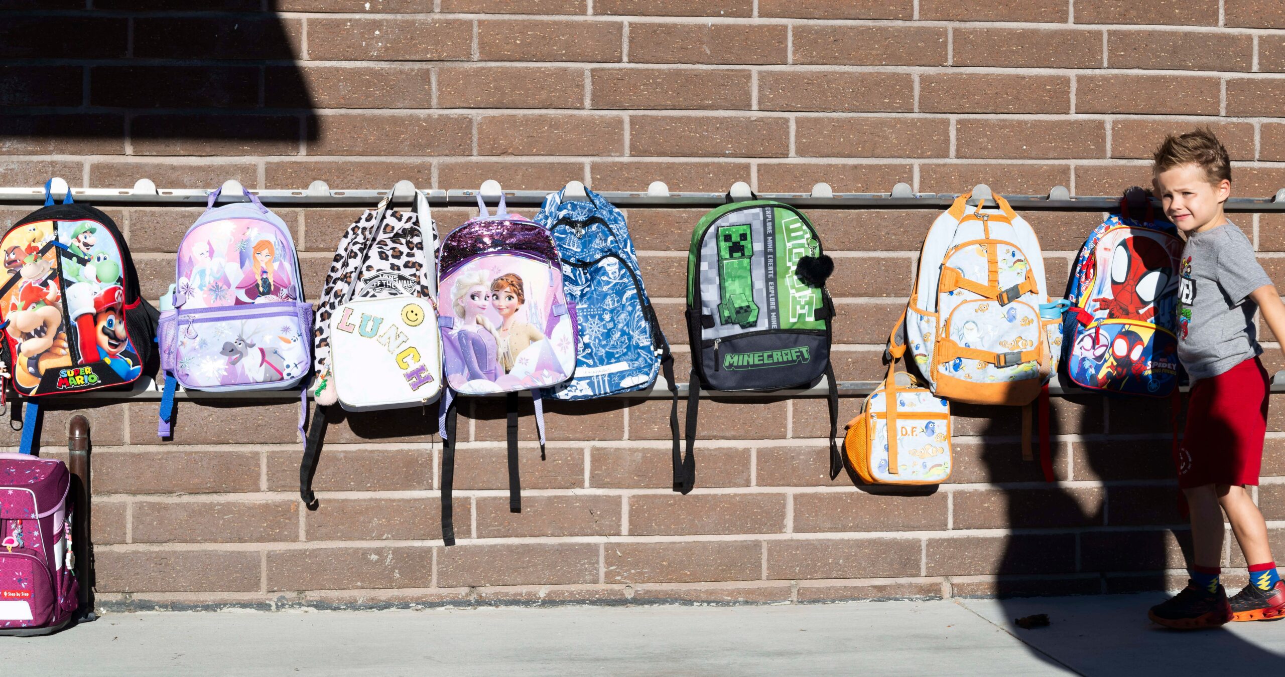 A young boy places a Spiderman backpack on a rack on a brick wall alongside other backpacks