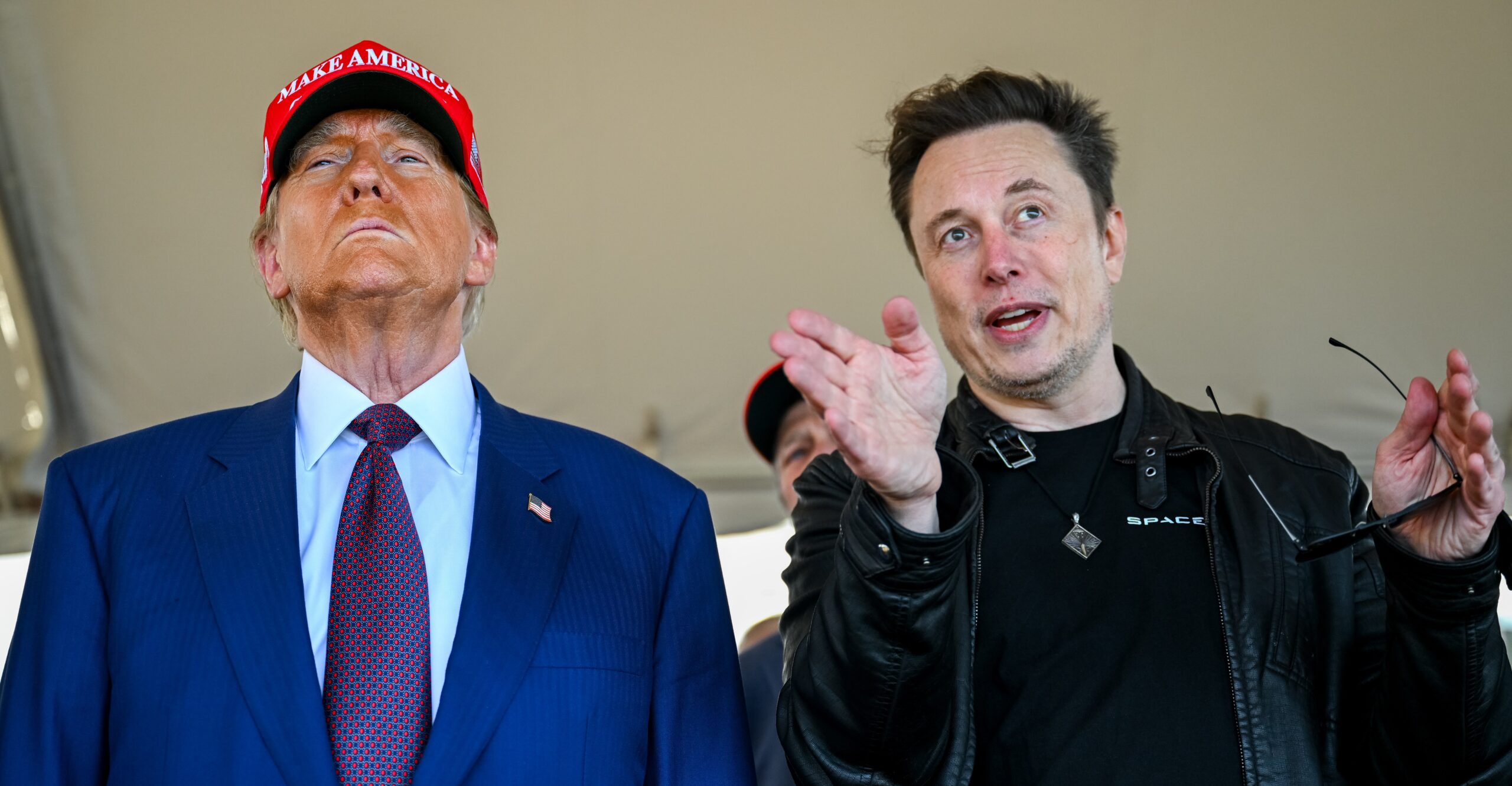 Trump looking ahead in a MAGA hat while Elon talks and gestures beside him.