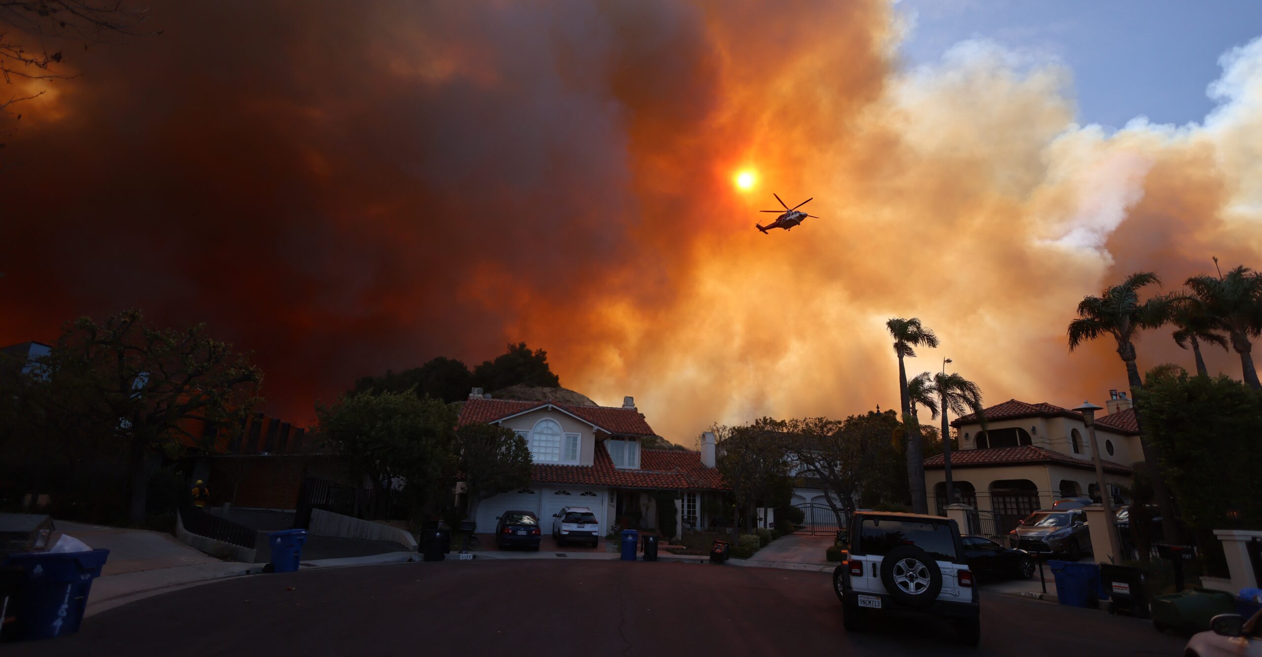 Expensive-looking homes at the end of a cul-de-sac. Behind them, the sky is saturated with smoke and an ominous orange color. A helicopter flies through it.