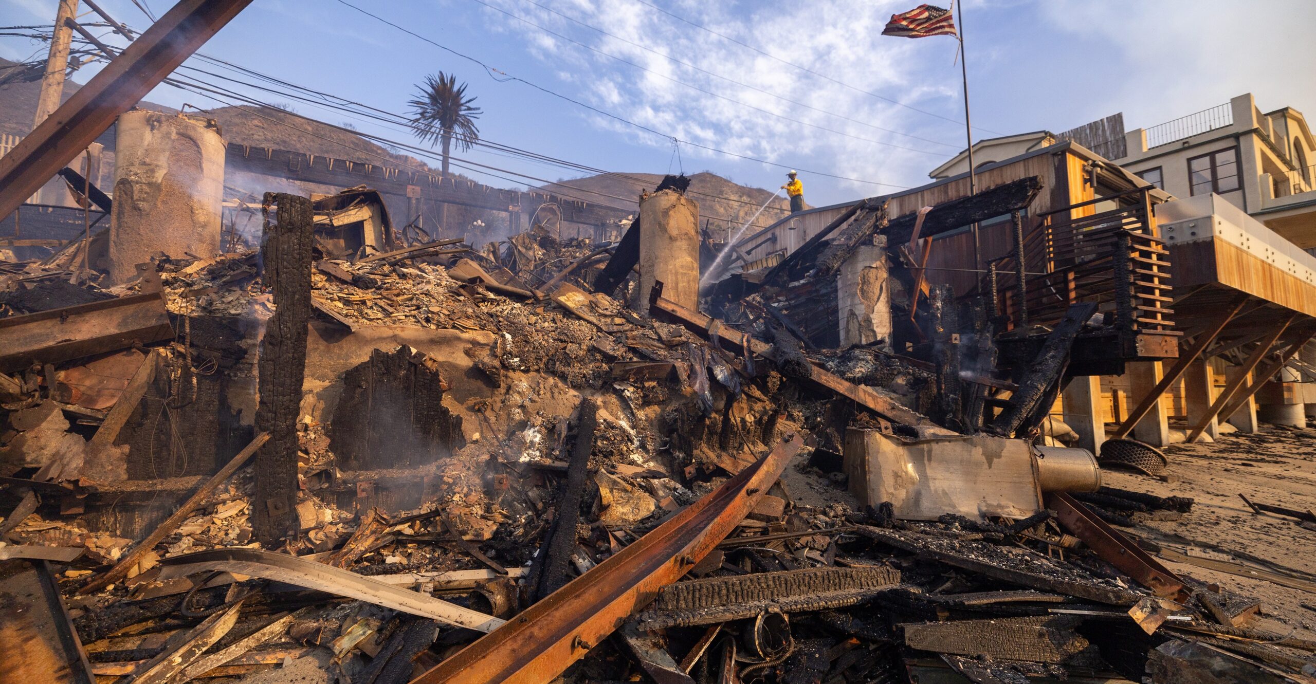 Seen from the beach below, a firefighter on a road above sprays down a pile of charred, twisted rubble, formerly a house on the beach.