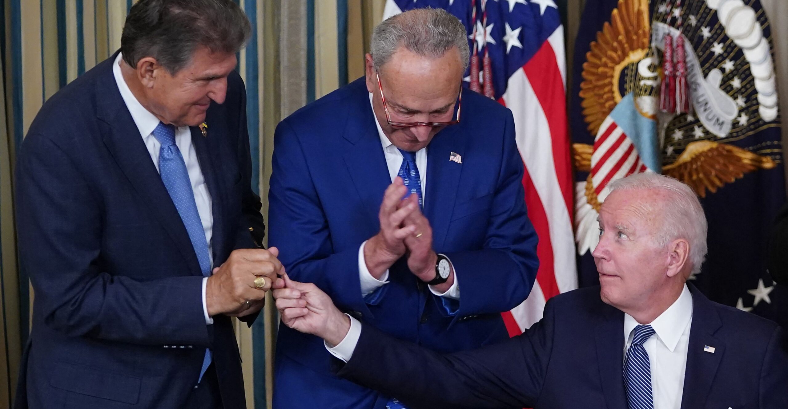 Biden, sitting down, hands Manchin, standing up, a pen 