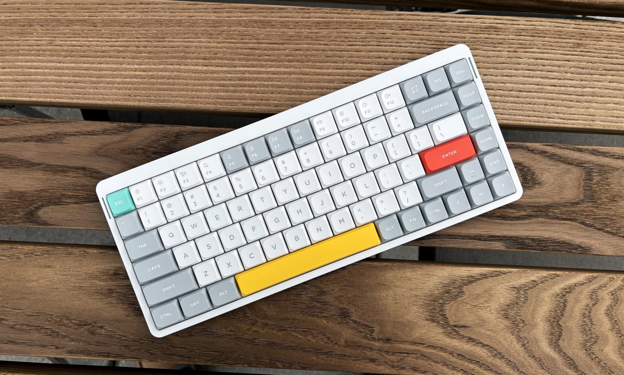 The NuPhy Air75 V2 mechanical keyboard rests on a brown wooden table. Its keys are mostly white and grey, with a yellow space bar, teal escape key and red enter key.