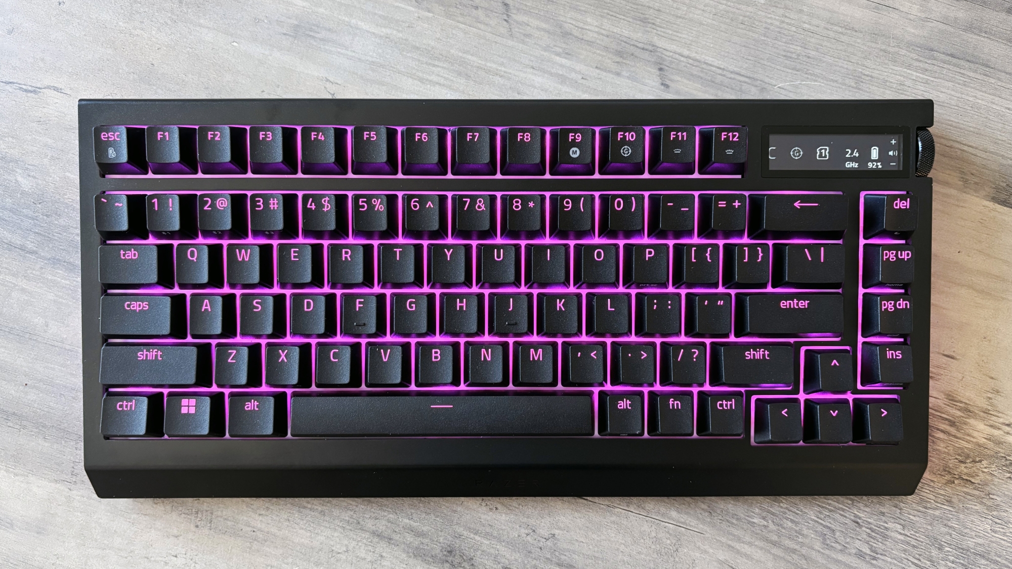 A black keyboard with pink RGB lighting and a black-and-white OLED display, the Razer BlackWidow V4 Pro 75%, rests atop a brown wooden table.