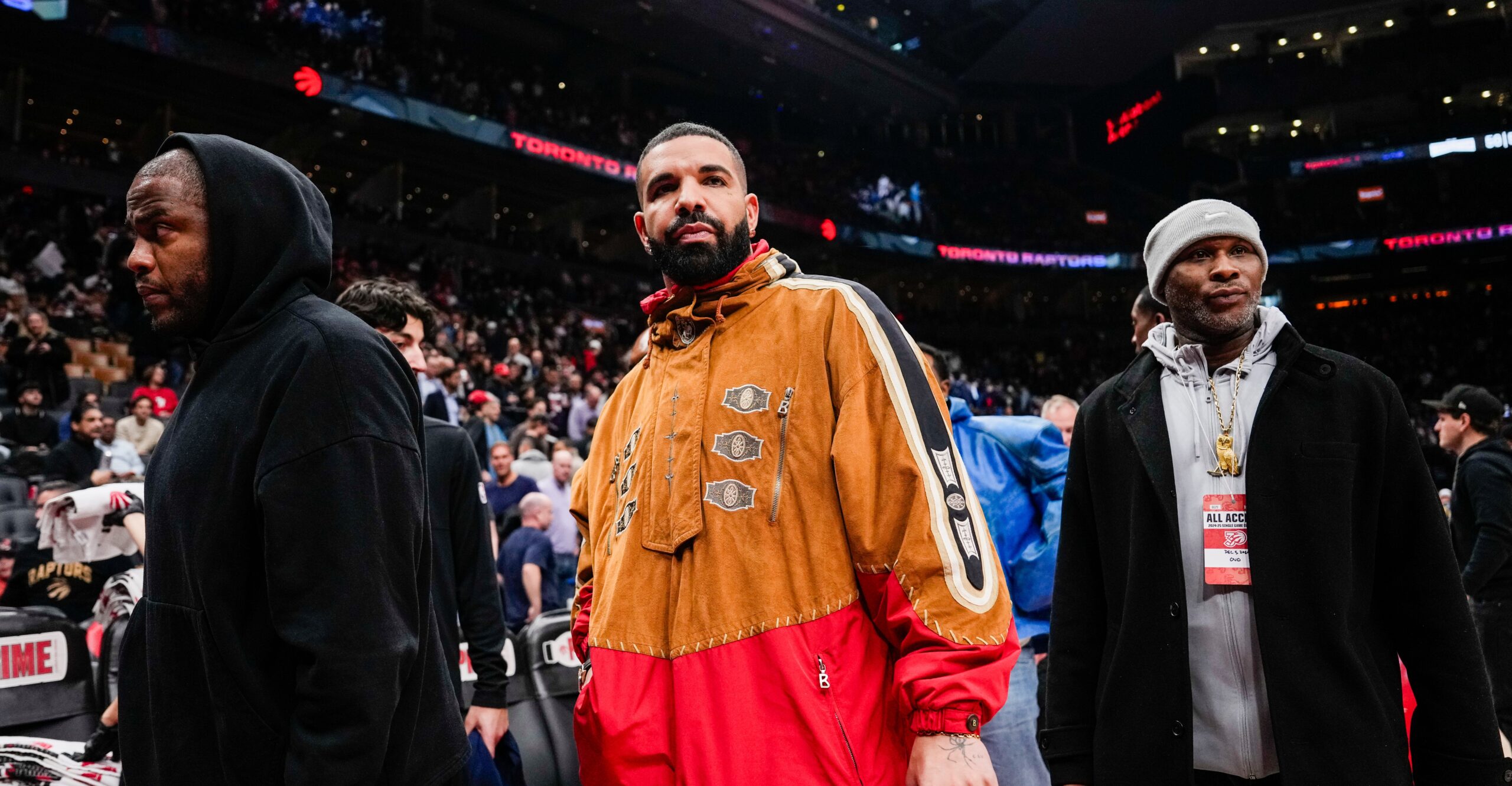 Drake wears an orange and red windbreaker with his bodyguards at a basketball game