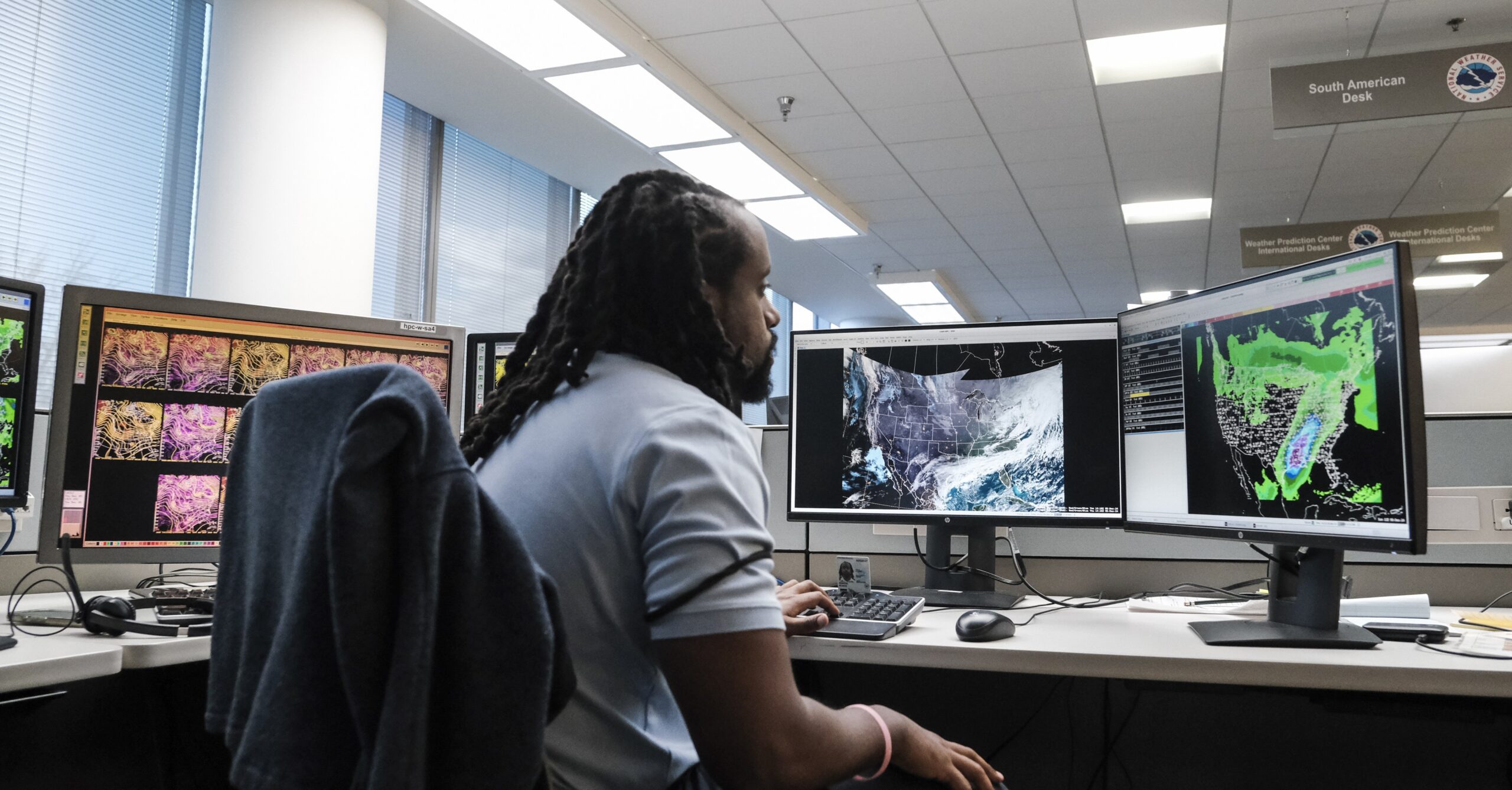 A man at a desk looks at several computer screens with maps of the US and weather blobs over the map.