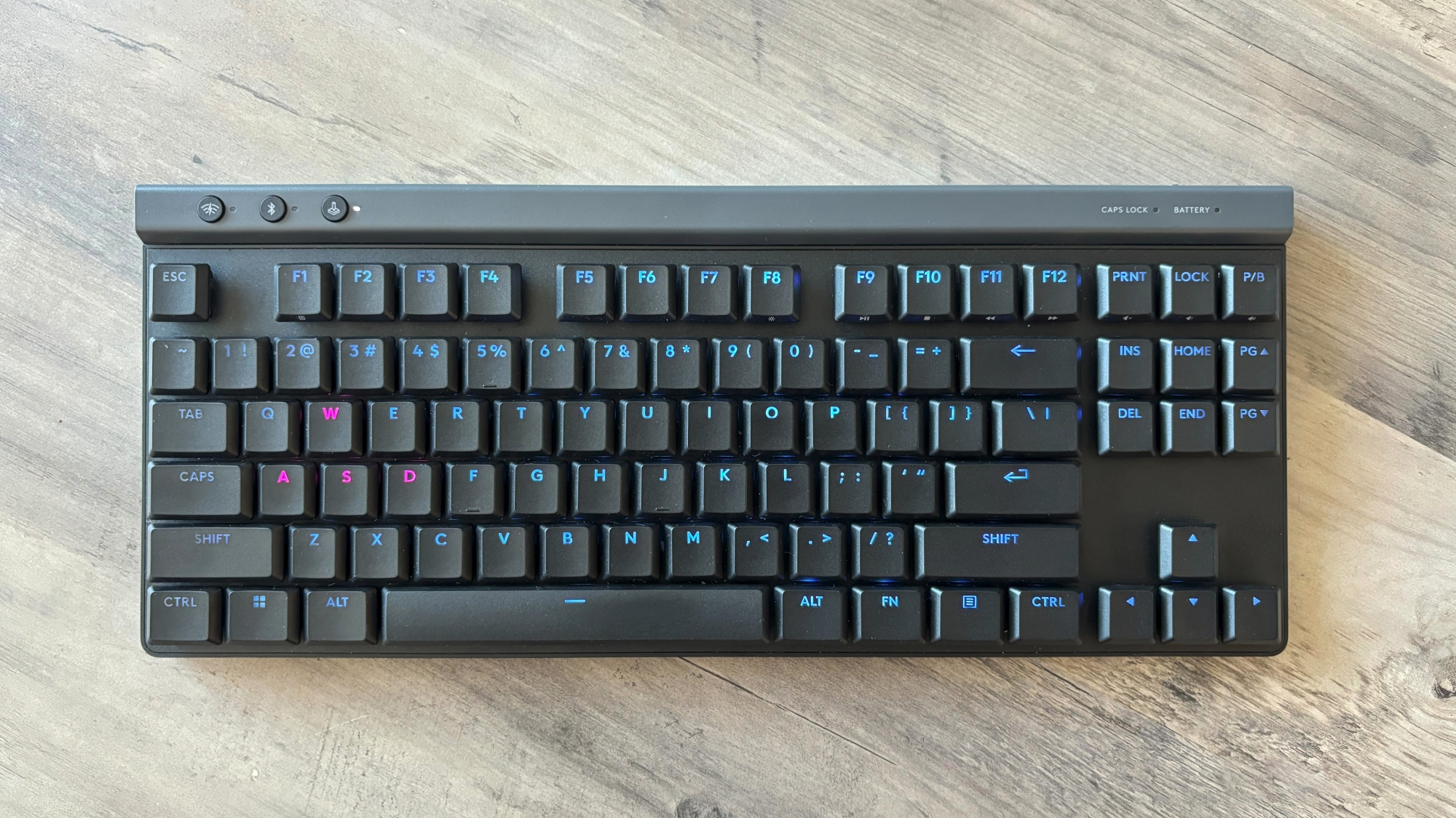 A black gaming keyboard with light blue RGB backlighting, the Logitech G515 TKL Lightspeed, rests atop a brown wooden table.