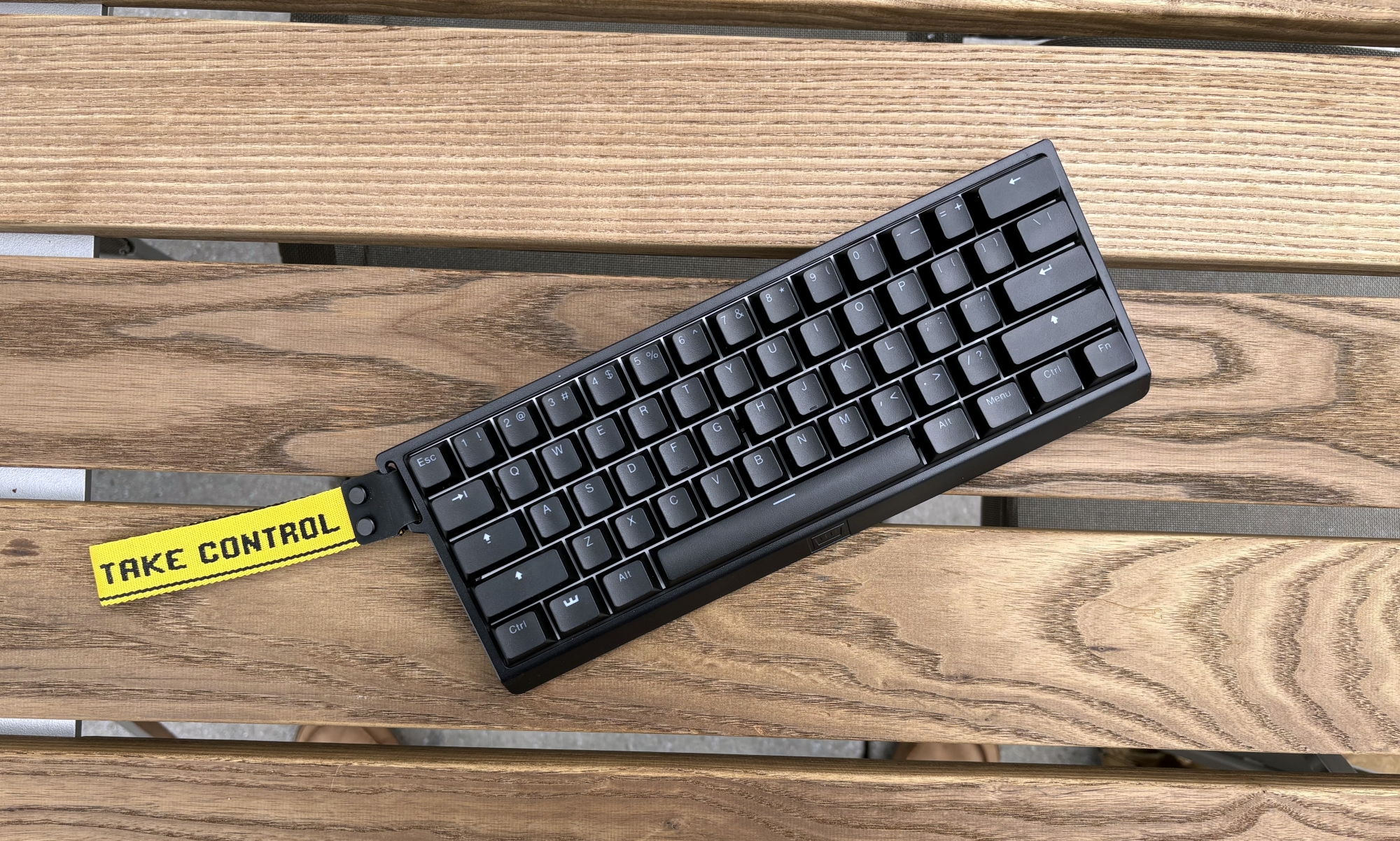 The Wooting 60HE+ gaming keyboard sits at an angle on a brown wooden outdoor table. The keyboard is all-black, but has a yellow ribbon attached to its top left corner, with the phrase 