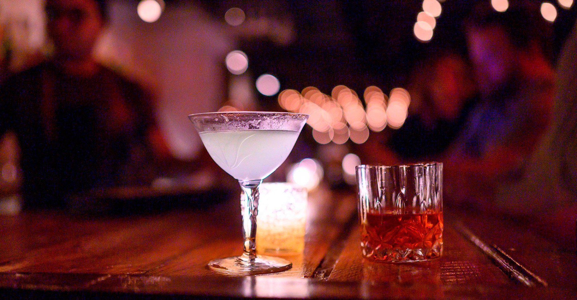 A blurry, dark bar scene with two cocktails in focus in the foreground