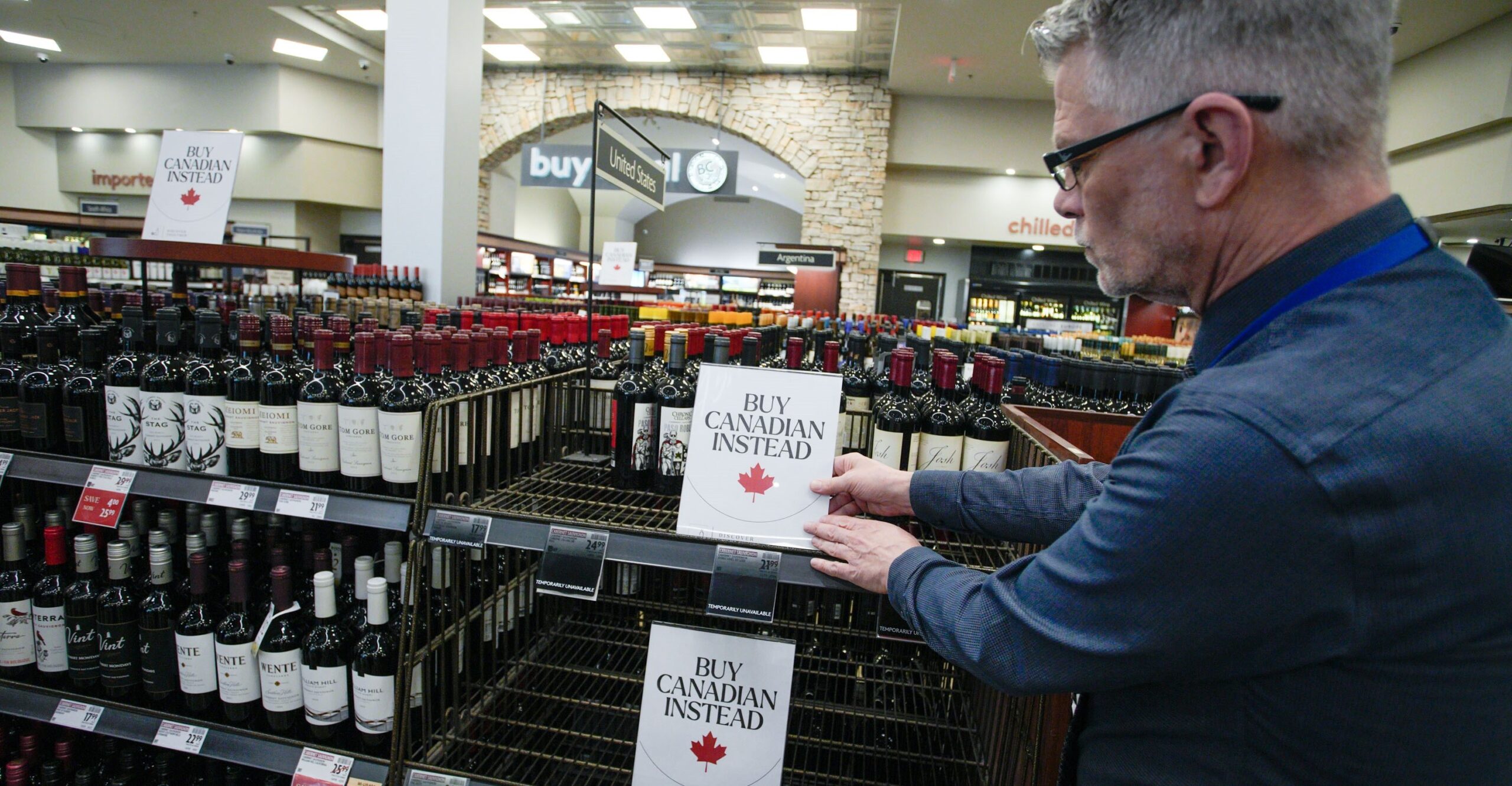 A grey haired man puts a sign with a red maple leaf on empty liquor store shelves that reads “Buy Canadian instead.”