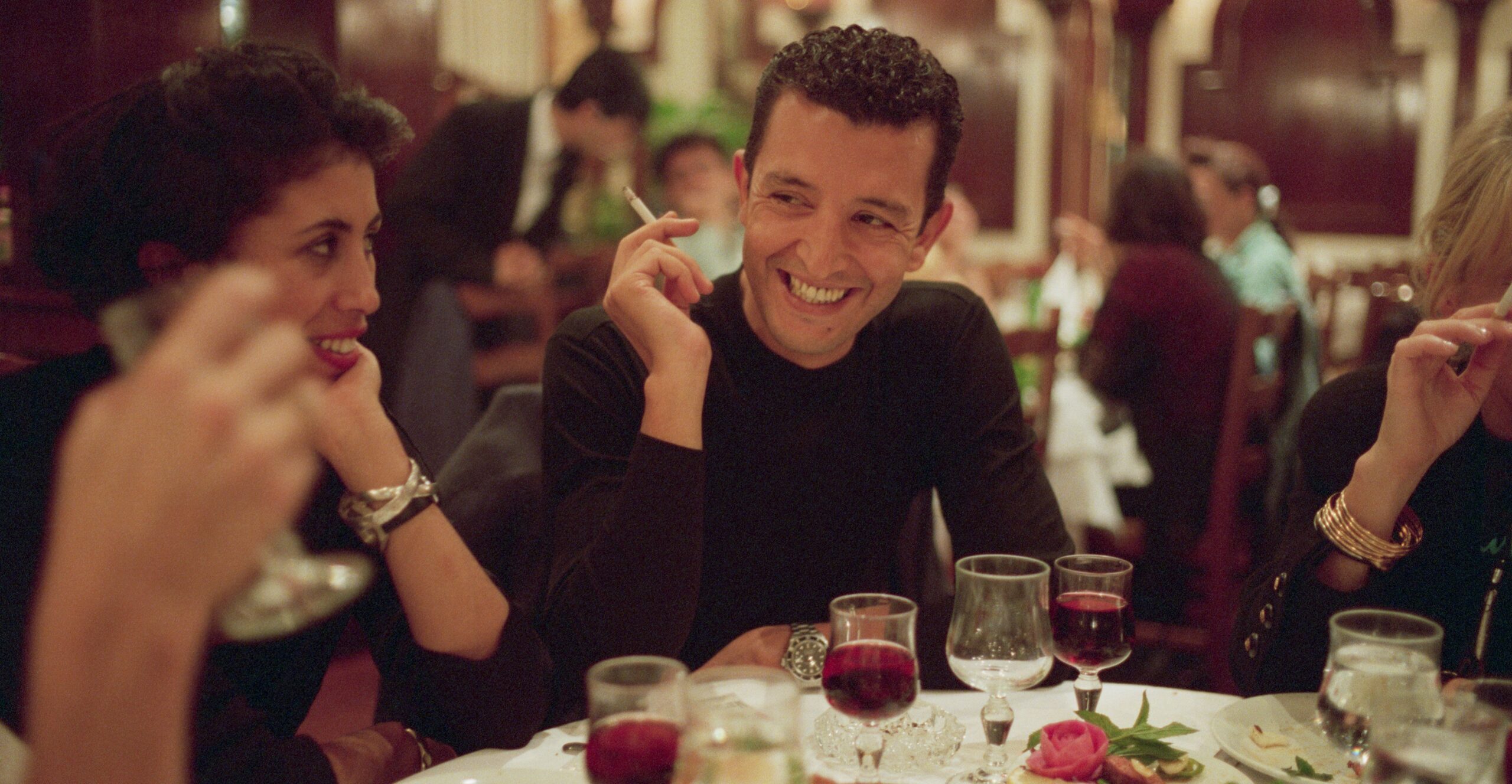 Man and women dining at a lively restaurant with food and red wine in front of them.