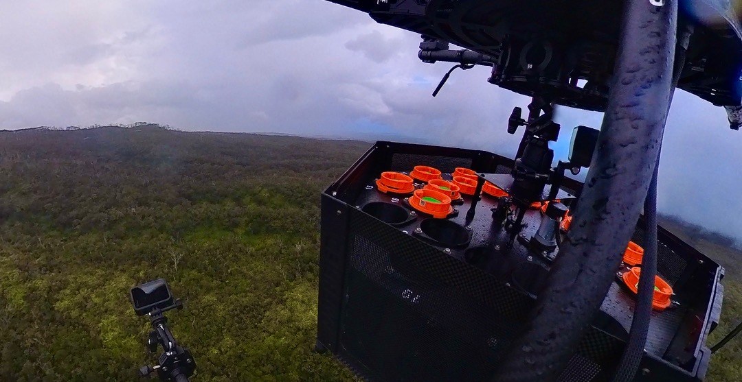 A drone’s eye view of its own frame, with a black box containing orange capsules attached to it, hovering over a forest that stretches out green to the horizon.