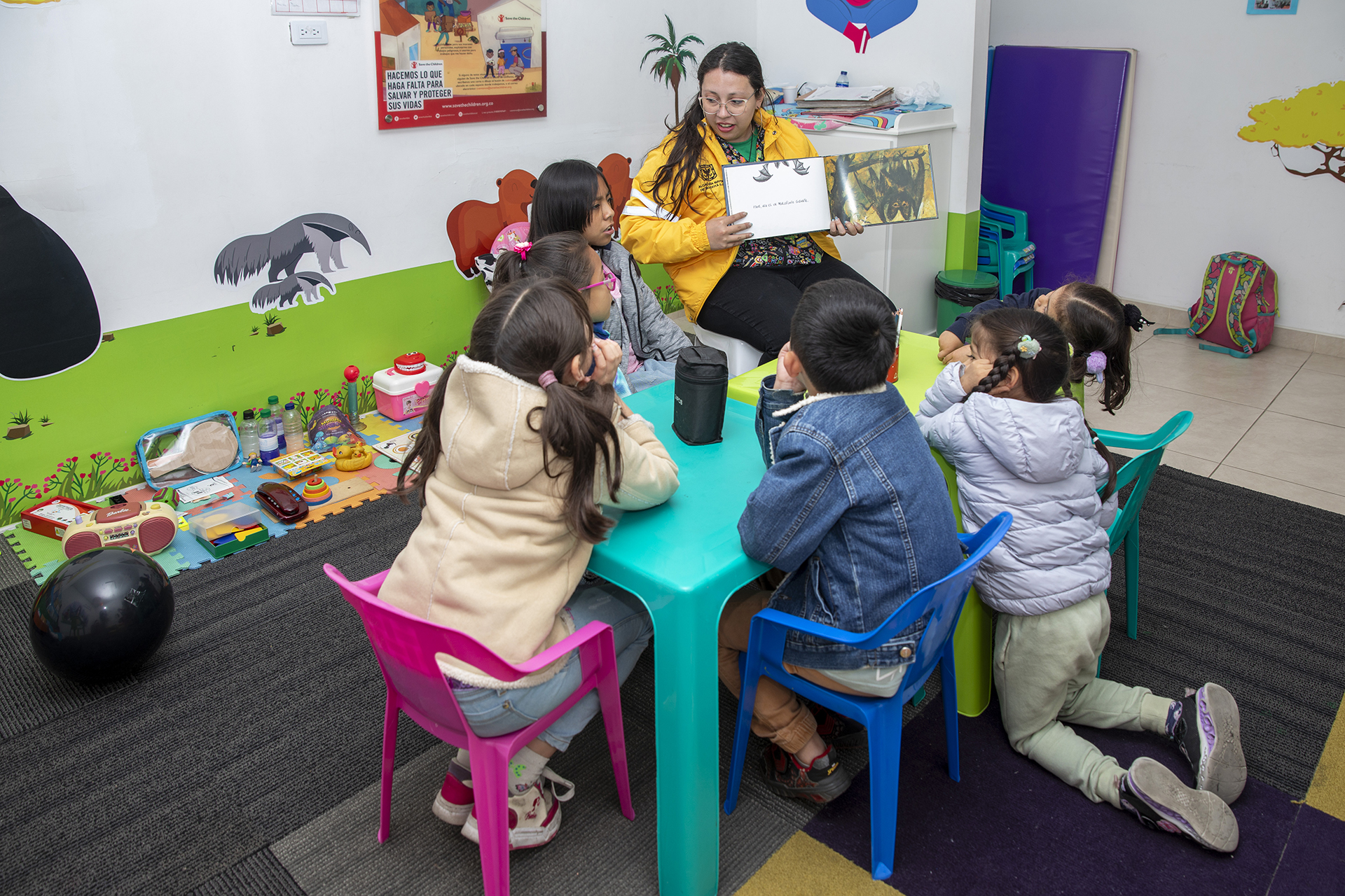 A young woman reads a story to six children of various ages at a small turquoise table in a classroom