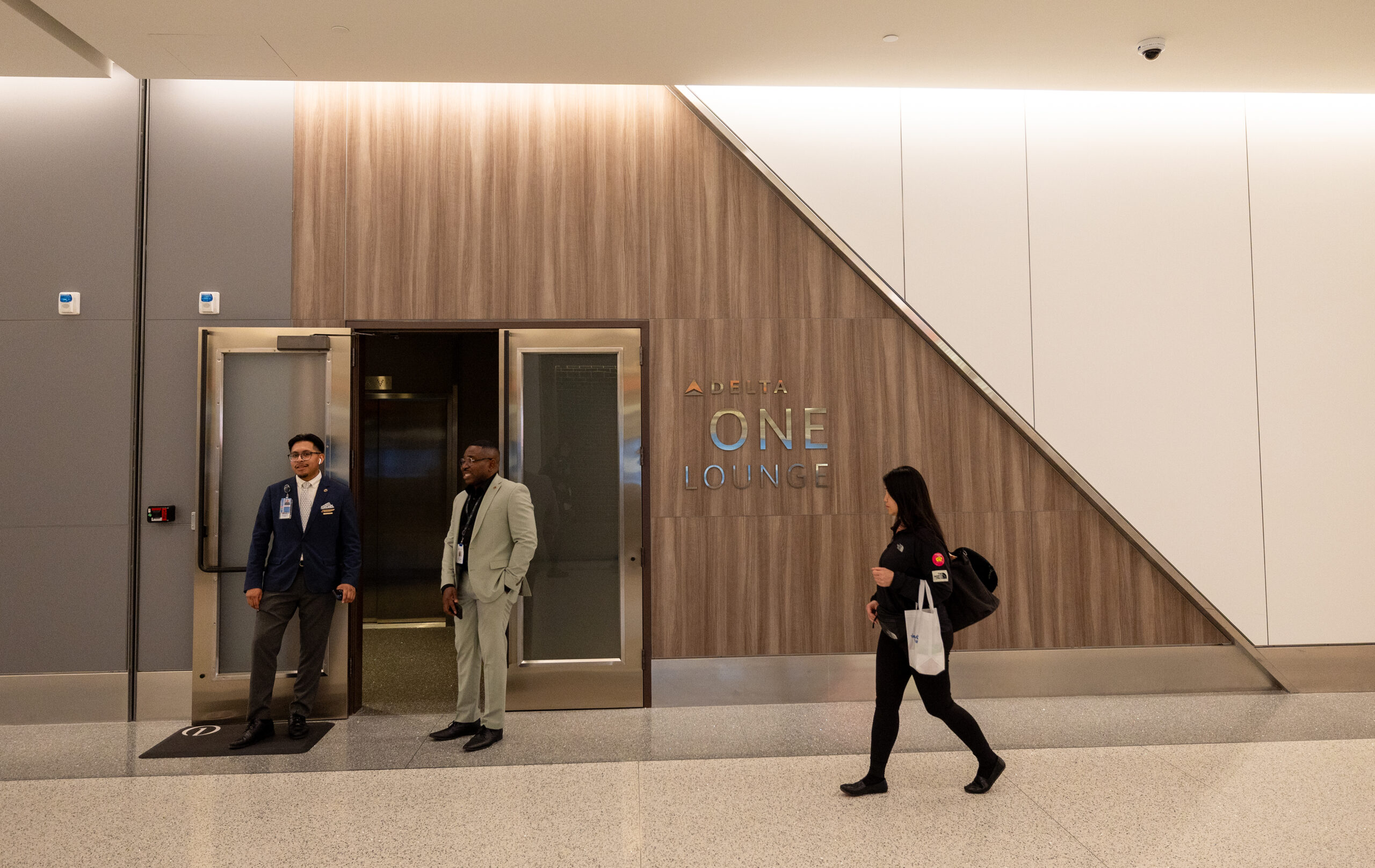 Two men stand in front of a lounge door while a woman walks by