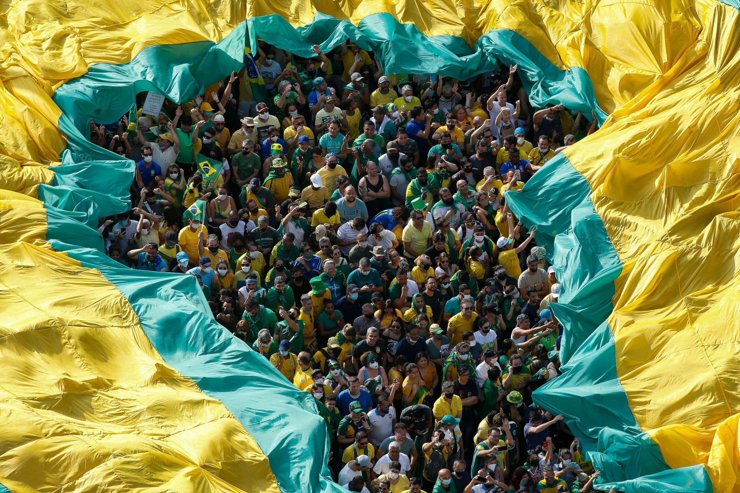 Overhead view of a large crowd dressed in green and yellow gathered beneath a massive piece of fabric in the colors of the Brazilian flag, which is stretched above them with an opening in the center revealing the people below