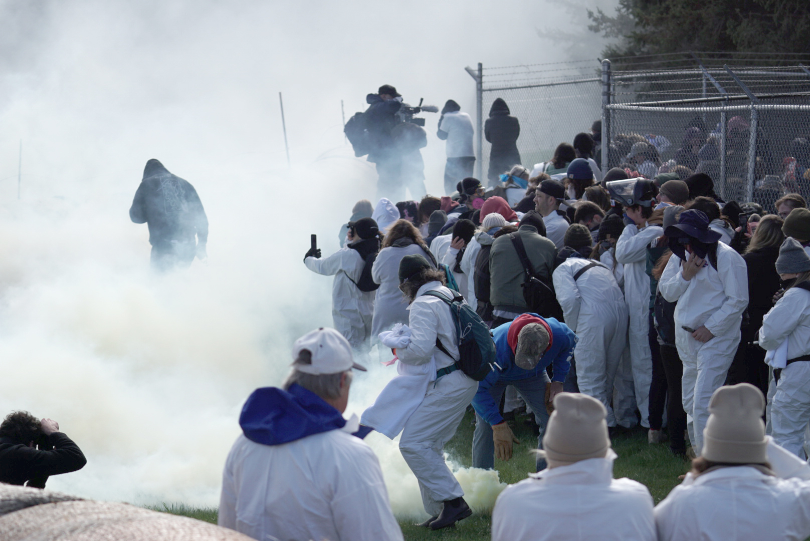 A large crowd of activists, many wearing white biosuits, huddle along a chain-link fence as thick tear gas fills the grassy area around them. Several people cover their faces or bend over in the smoke, while others film with phones and a camera crew stands in the haze.
