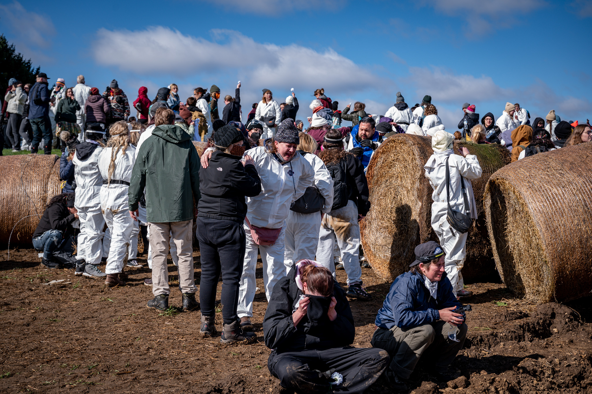 A large crowd of activists, many wearing white biosuits, gathers around rows of hay bales in a field under a blue sky. In the foreground, one person sits on the ground covering their face while others nearby talk, crouch, or help each other.