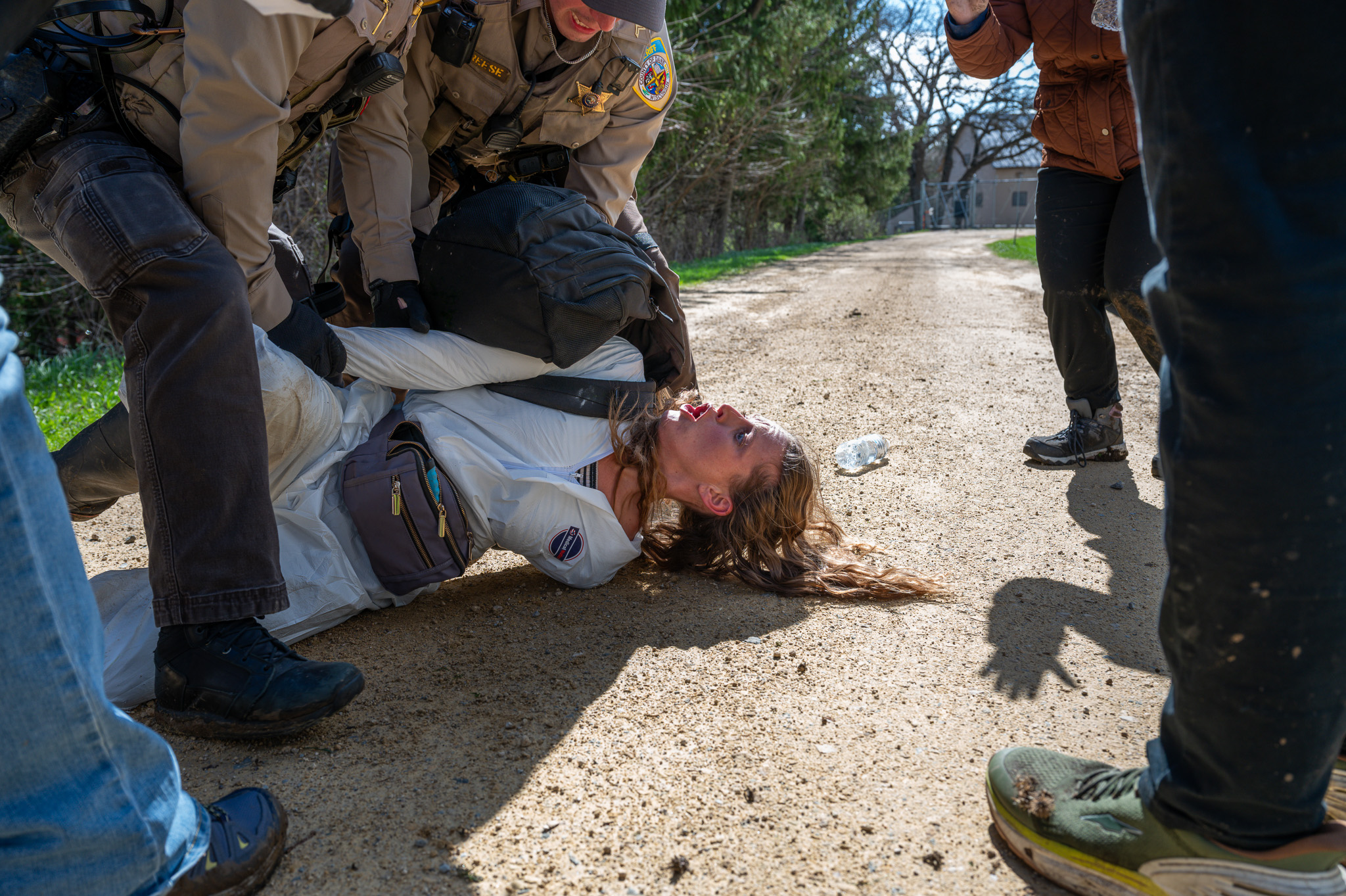 A woman in white biosuit lies on her back on a gravel road, mouth open, as two uniformed law enforcement officers kneel over and restrain her. Bystanders stand close by, and a plastic water bottle lies on the ground nearby.