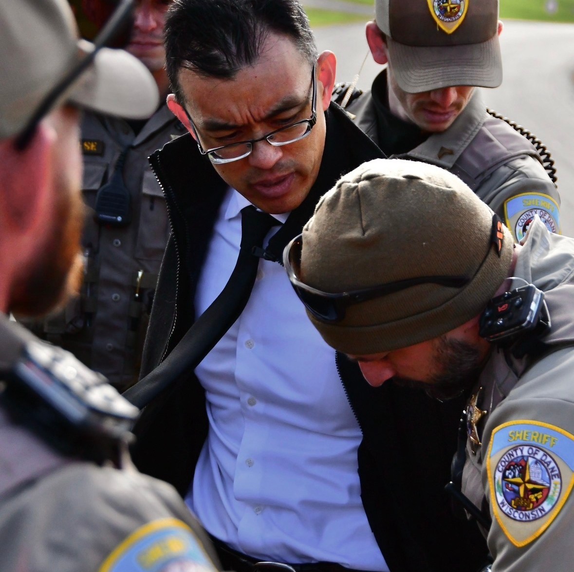Dane County sheriff’s deputies surround and restrain a man in glasses, a white shirt, black tie, and black jacket during an outdoor protest.