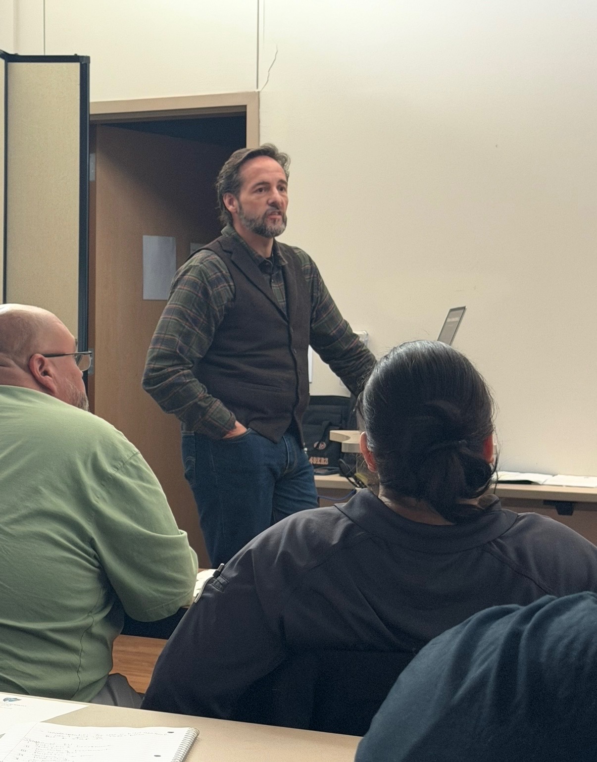 A man stands in front of a classroom talking. You can see the backs of two students’ heads. 