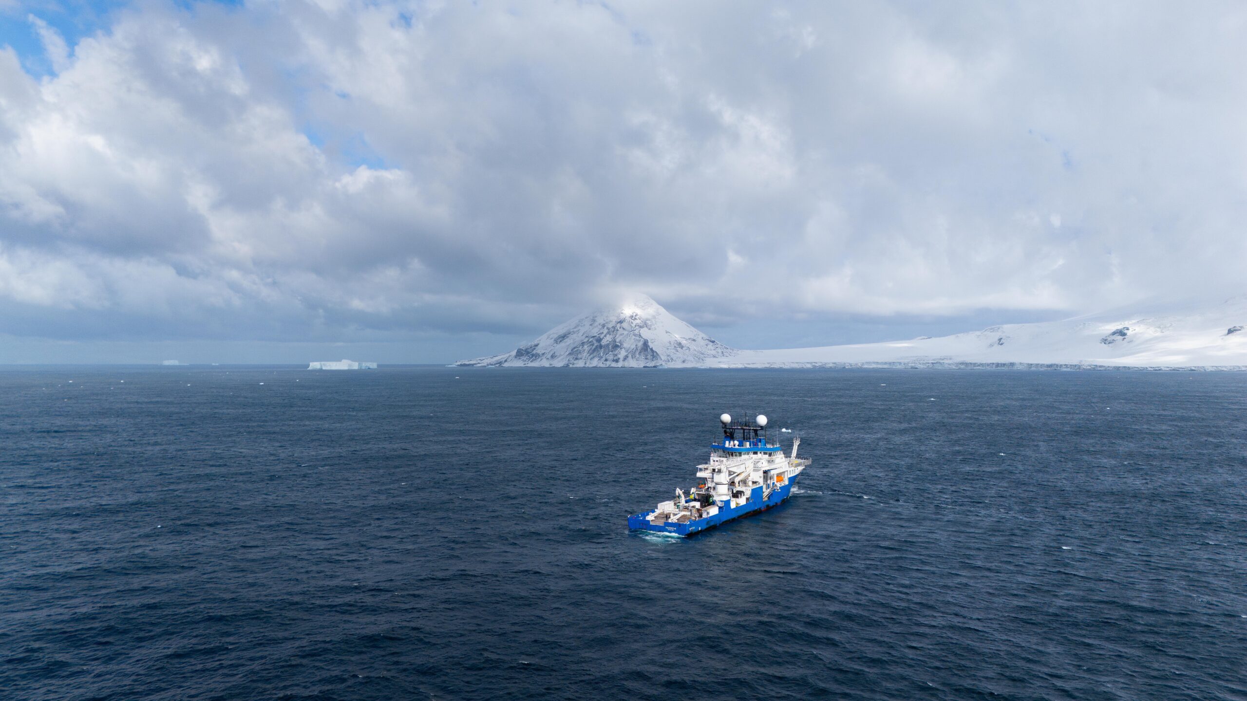A research vessel, with a blue hull and lots of white research equipment on deck, sails on a dark sea with snow-covered shores and mountains in the background.