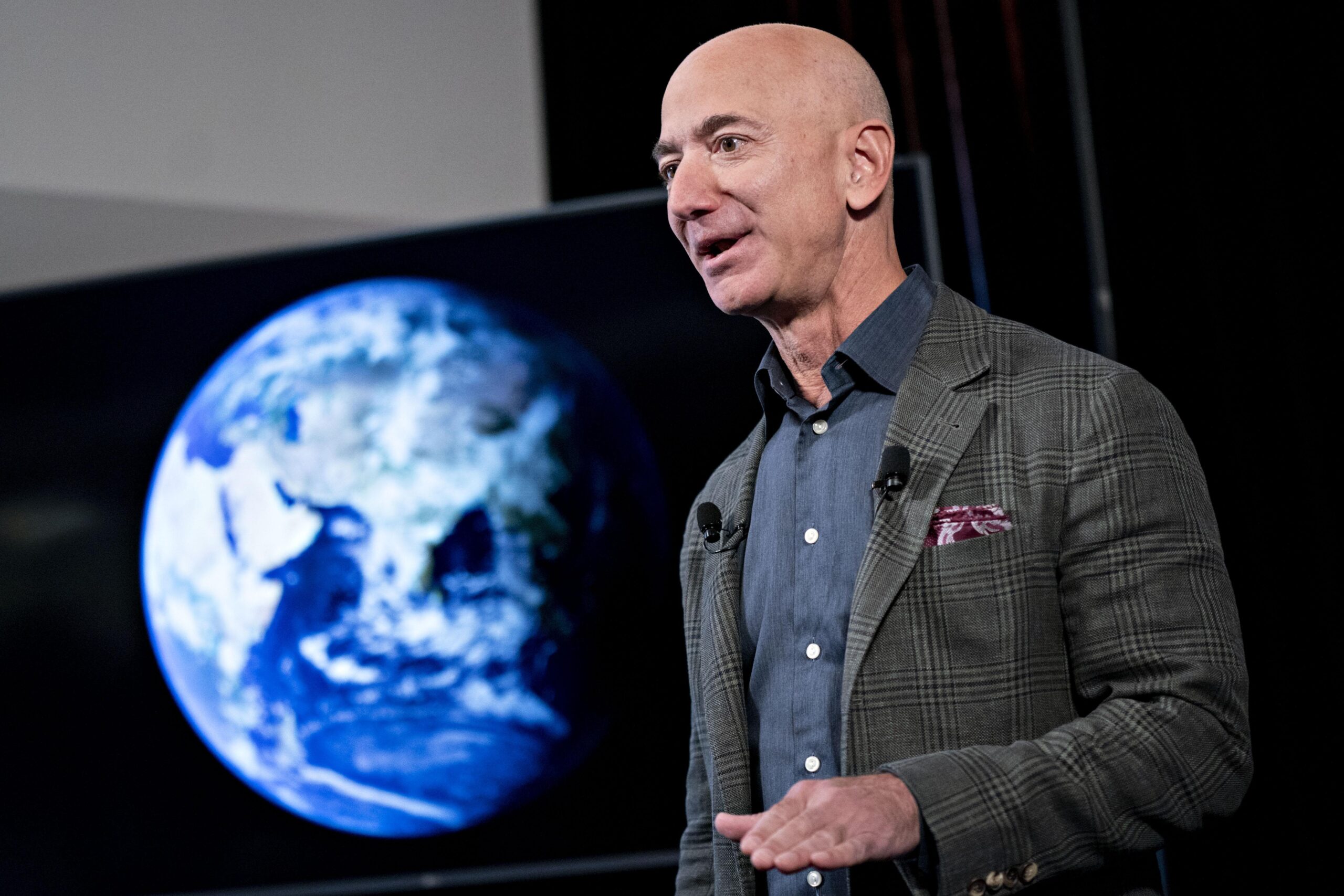 Jeff Bezos, in a gray jacket and blue shirt, standing near a large screen showing the planet Earth.