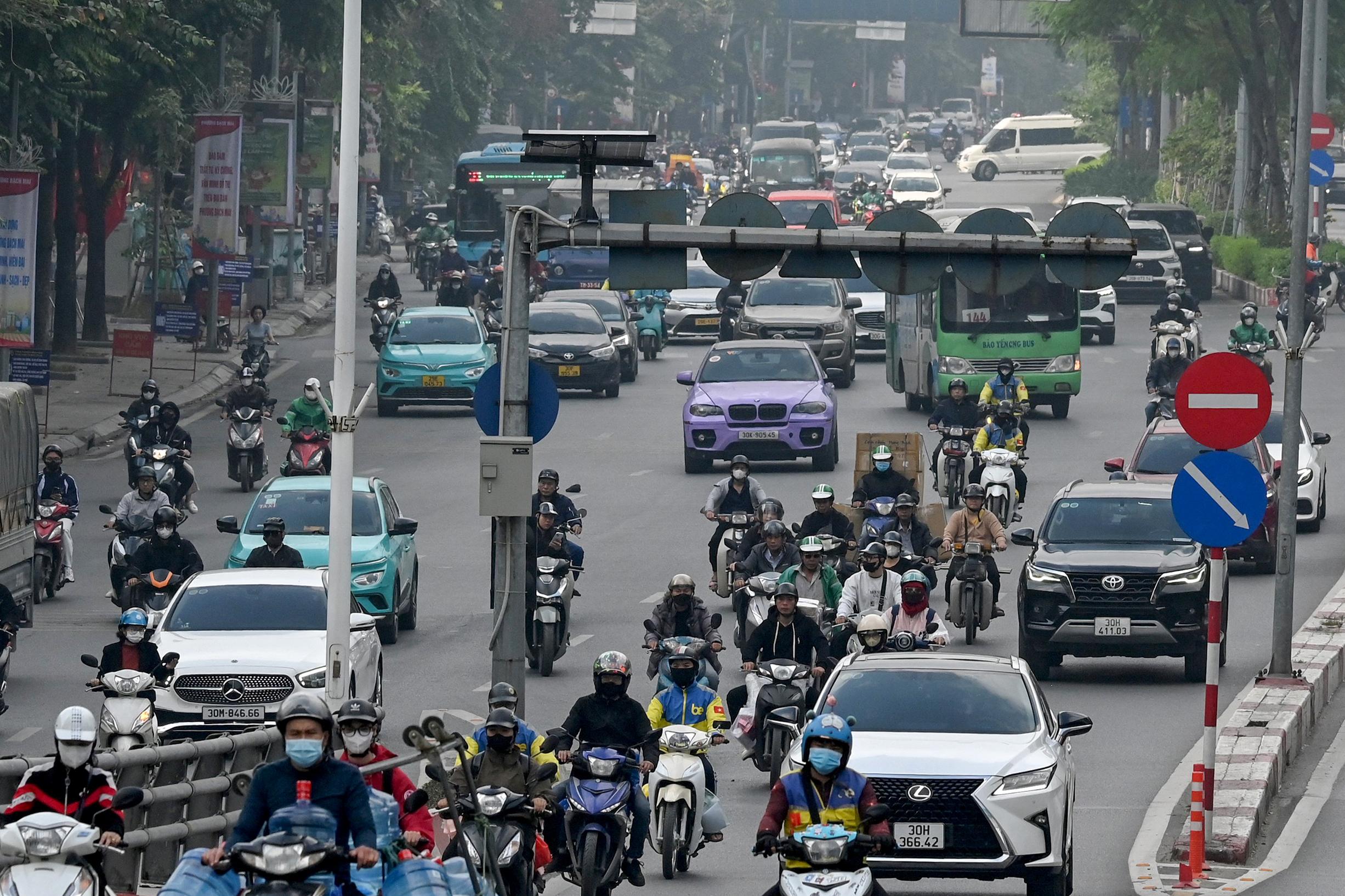 Dense urban traffic of motorbikes, cars, taxis, and buses fills a hazy multilane street, with riders packed closely together in mixed traffic.