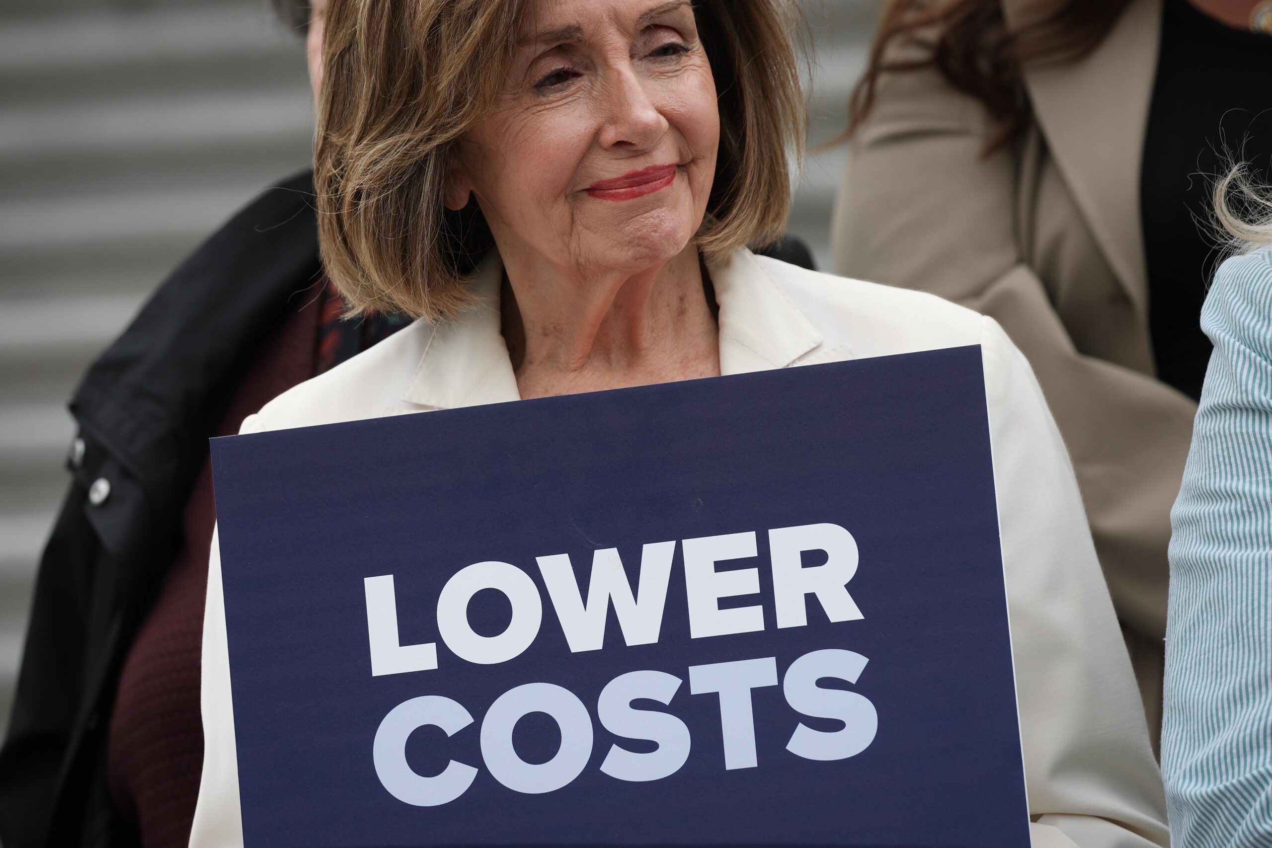 Pelosi holding a sign that says “lower costs”