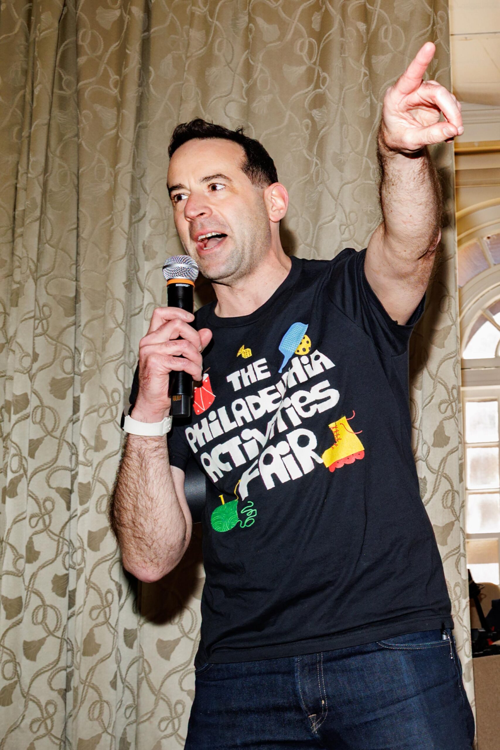 a man animatedly speaks into a microphone while pointing out of frame. His shirt reads “The Philadelphia Activities Fair”