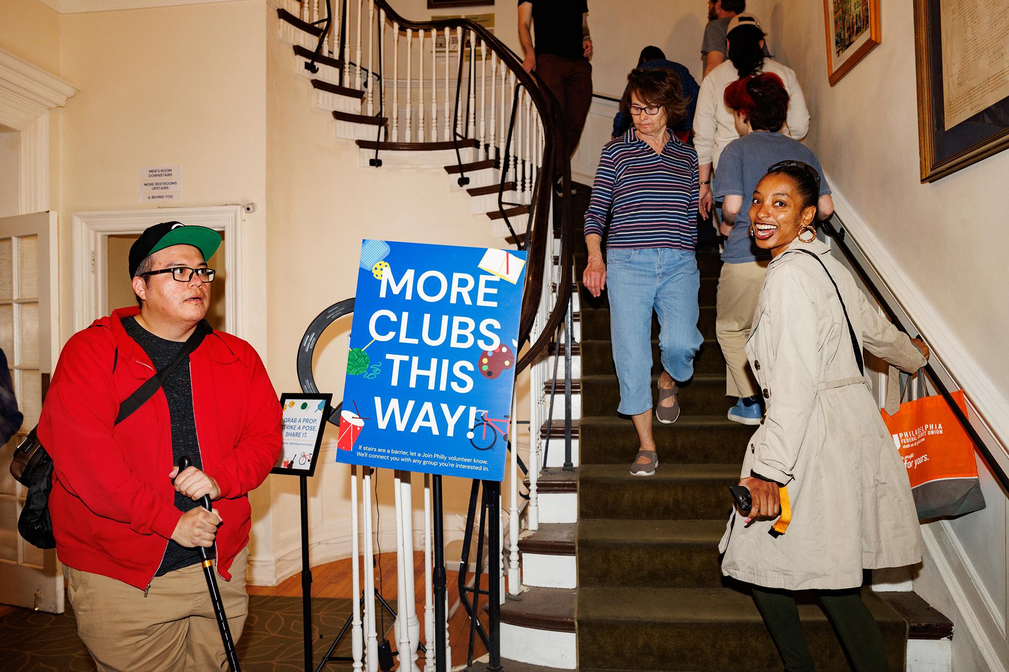 a woman smiling at the camera as she ascents a staircase full of people. A nearby blue sign reads “More clubs this way!”