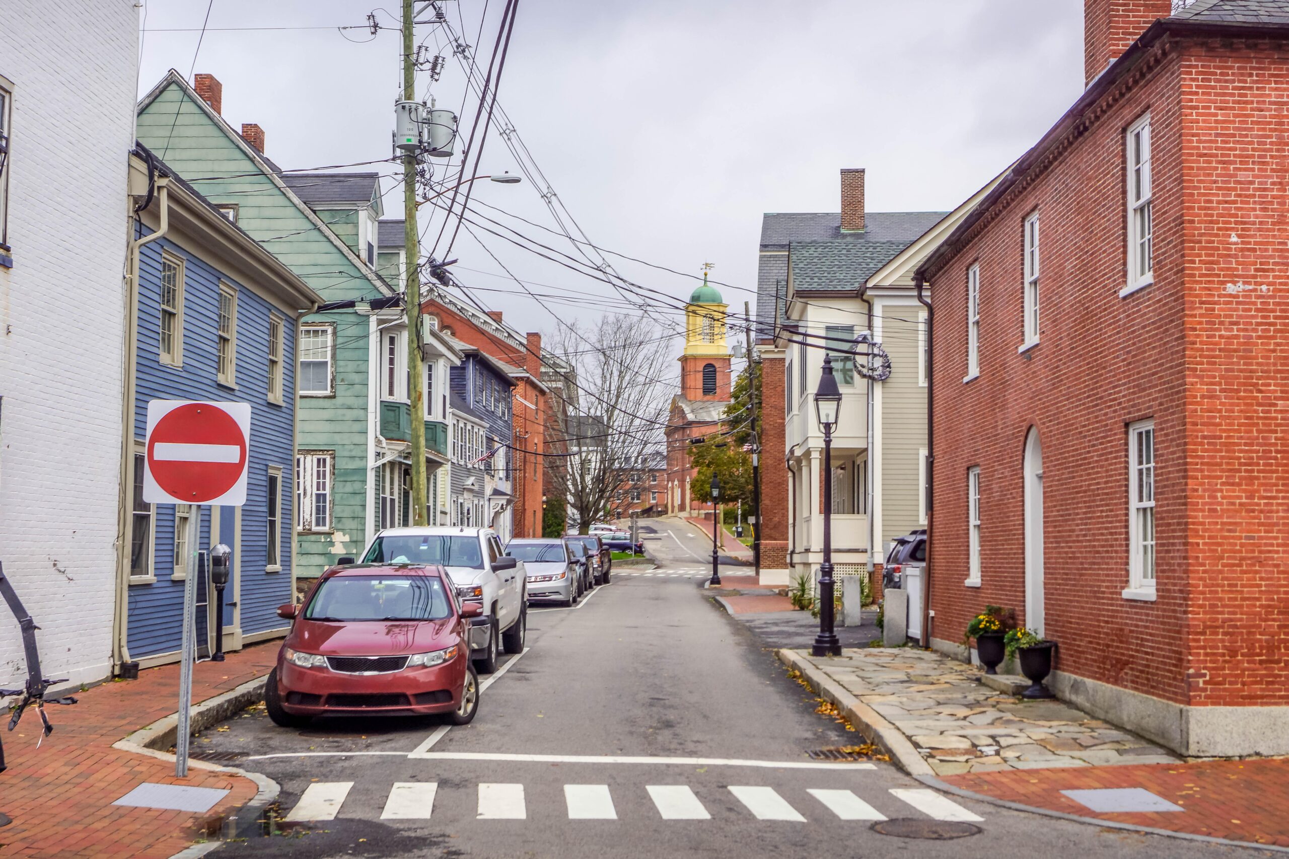 A narrow street in an older, built-up neighborhood lined with historic brick and clapboard buildings, parked cars, brick sidewalks, overhead utility wires, and a church tower in the background.