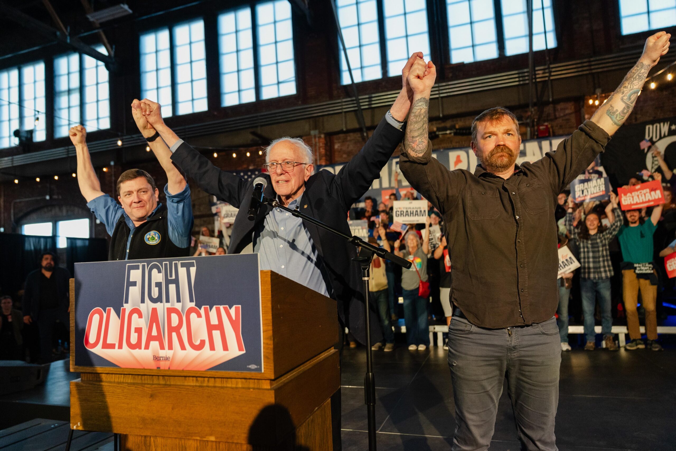 Sen. Bernie Sanders at a podium holding hands in a victory pose with Troy Jackson and Graham Platner.