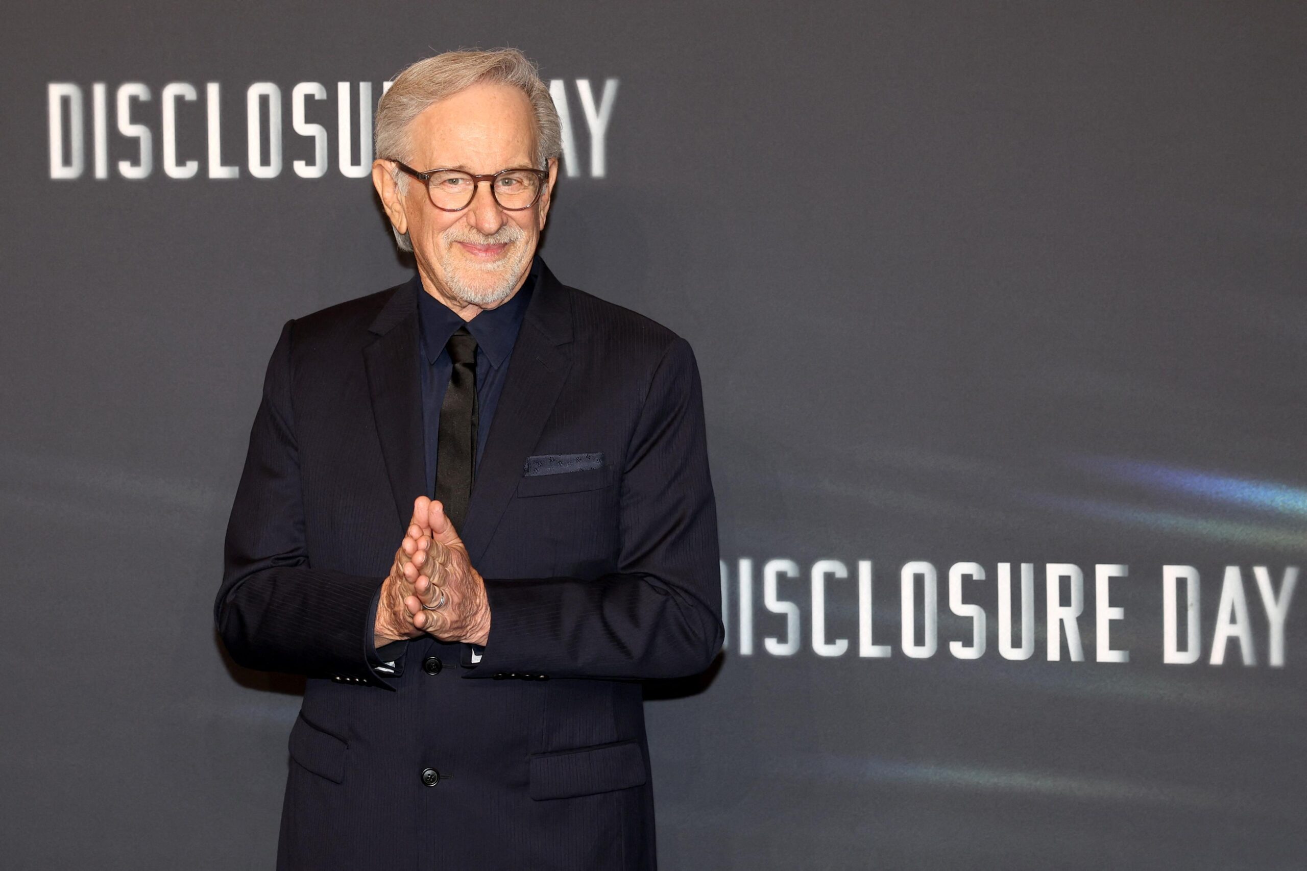 Steven Spielberg, in a black suit, stands in front of a backdrop reading “Disclosure Day.” Steven Spielberg, in a black suit, stands in front of a backdrop reading “Disclosure Day.”