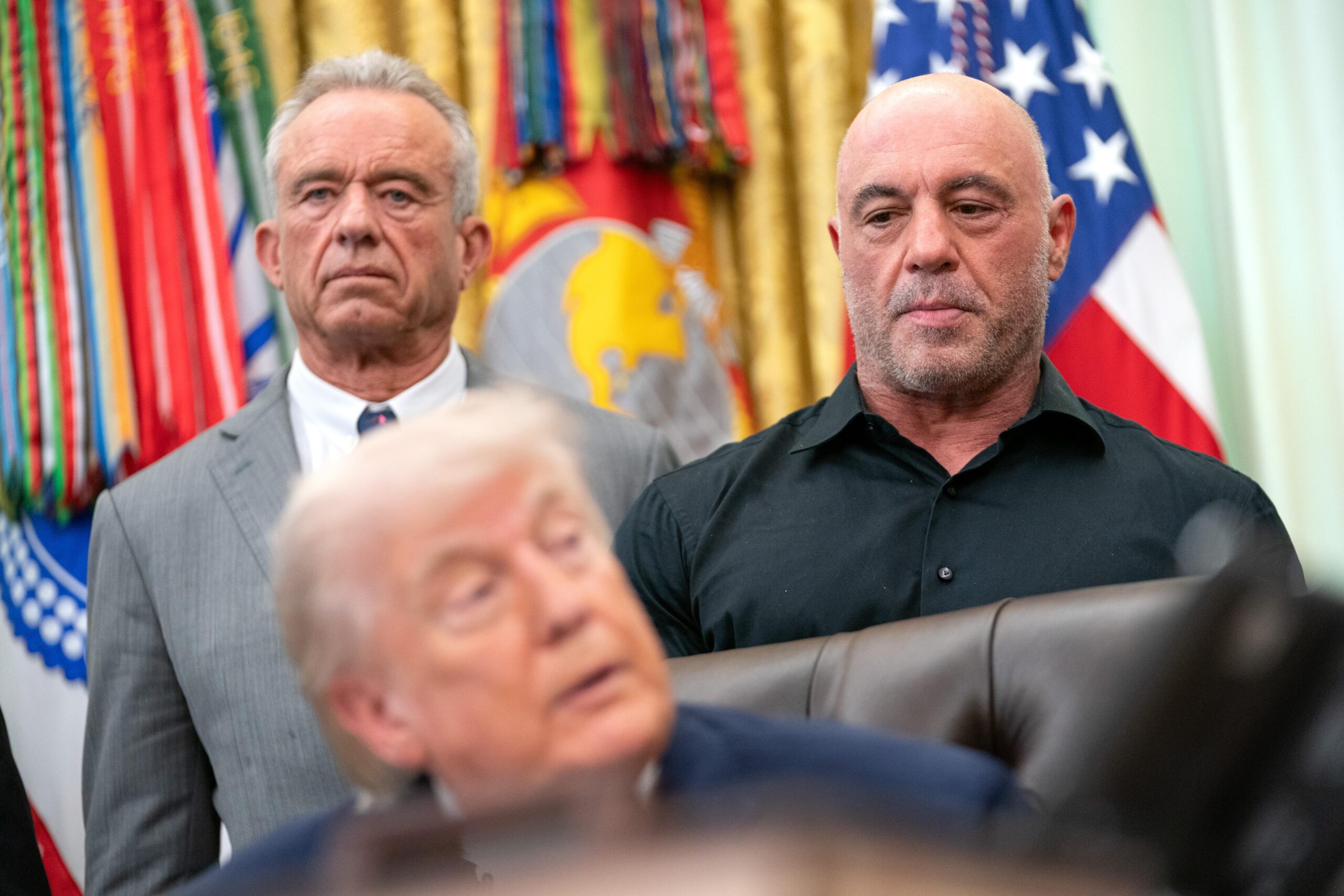 Trump is sitting in his chair and RFK Jr and Joe Rogan are standing behind him in the Oval Office
