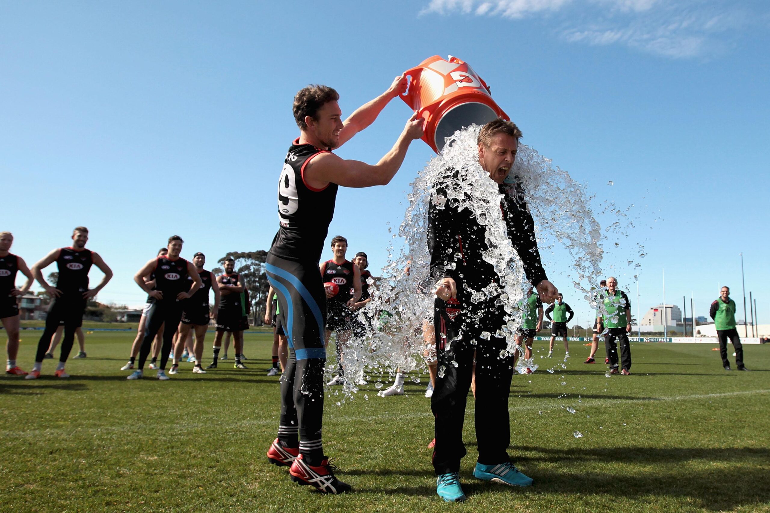 A man pours a bucket of ice water over another man while standing on a sports field
