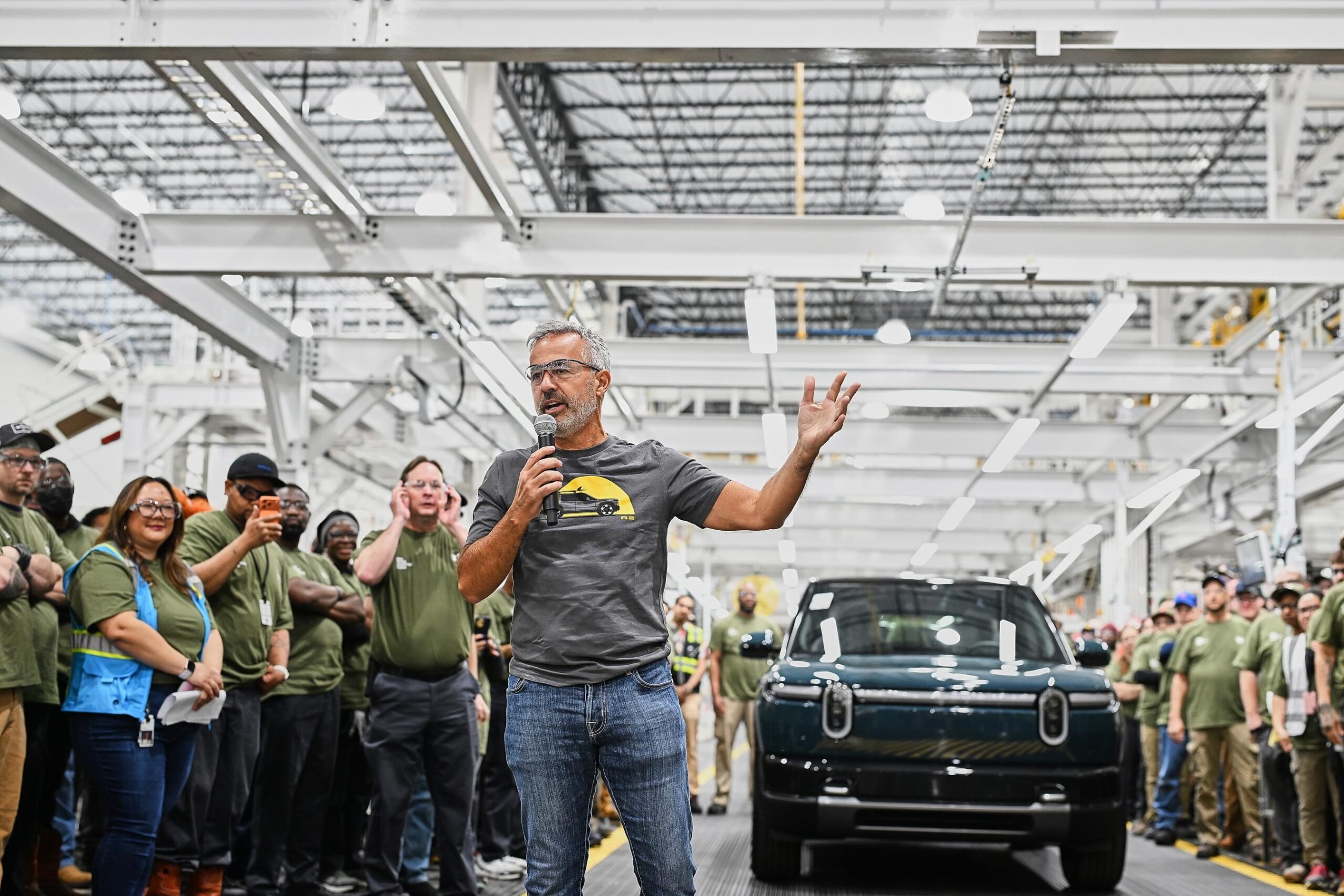 A man in a grey t-shirt and jeans holds a microphone and stands in front of a blue Rivian r2 inside of a huge, bright factory space and speaks to a crowd of people who are mostly wearing green t-shirts. A man in a grey t-shirt and jeans holds a microphone and stands in front of a blue Rivian r2 inside of a huge, bright factory space and speaks to a crowd of people who are mostly wearing green t-shirts.